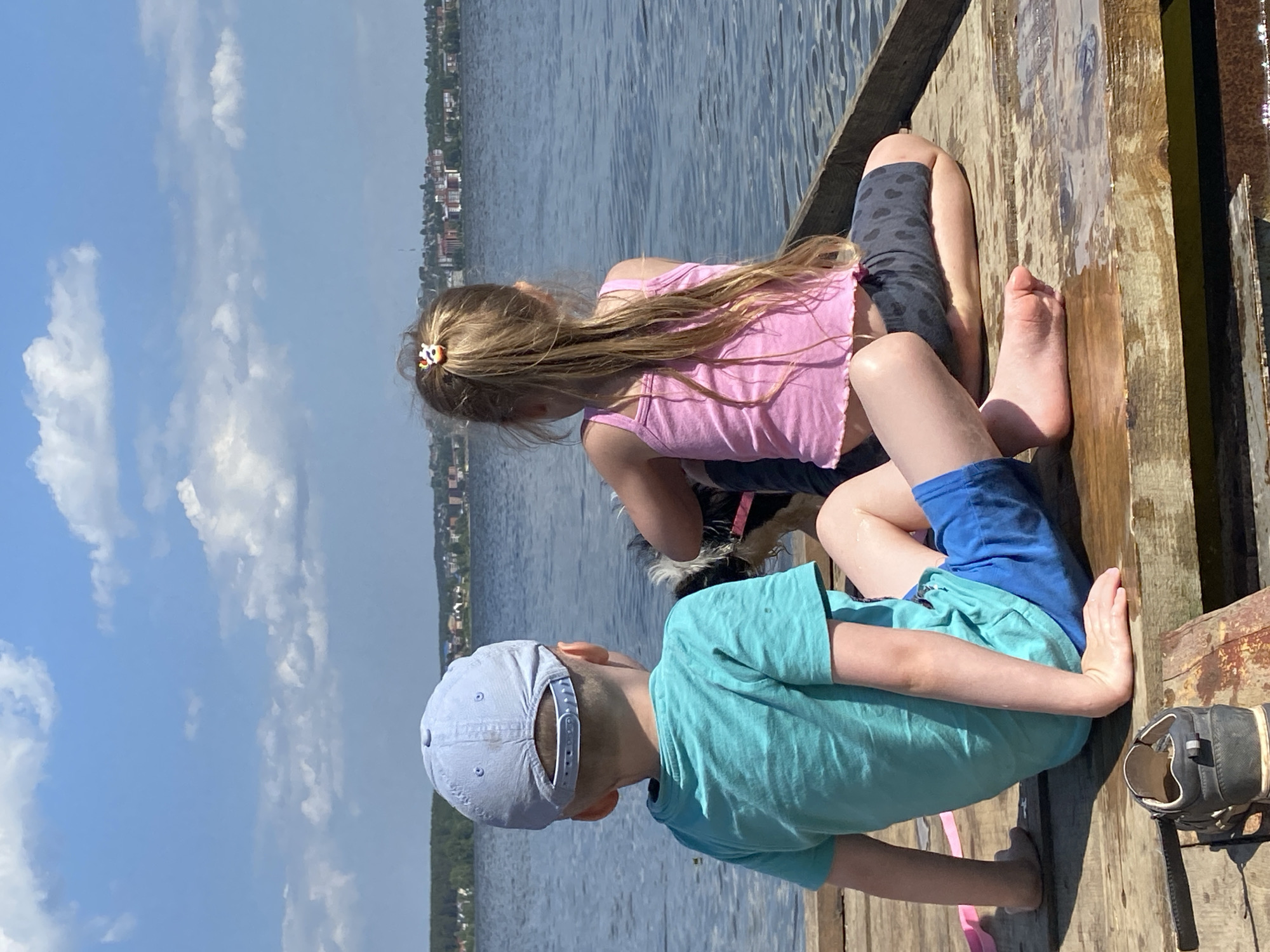 Two children are sitting on a wooden dock, facing the water. A girl with long blonde hair is wearing a pink top and dark leggings. A boy wearing a blue shirt and shorts sits beside her, his back to the camera. Both children are barefoot. A dog is visible between them. The location appears to be a body of water, possibly in the city of Votkinsk, Russia, suggested by the buildings in the background. The sky is blue with scattered clouds. A pair of shoes lies on the dock near the boy.