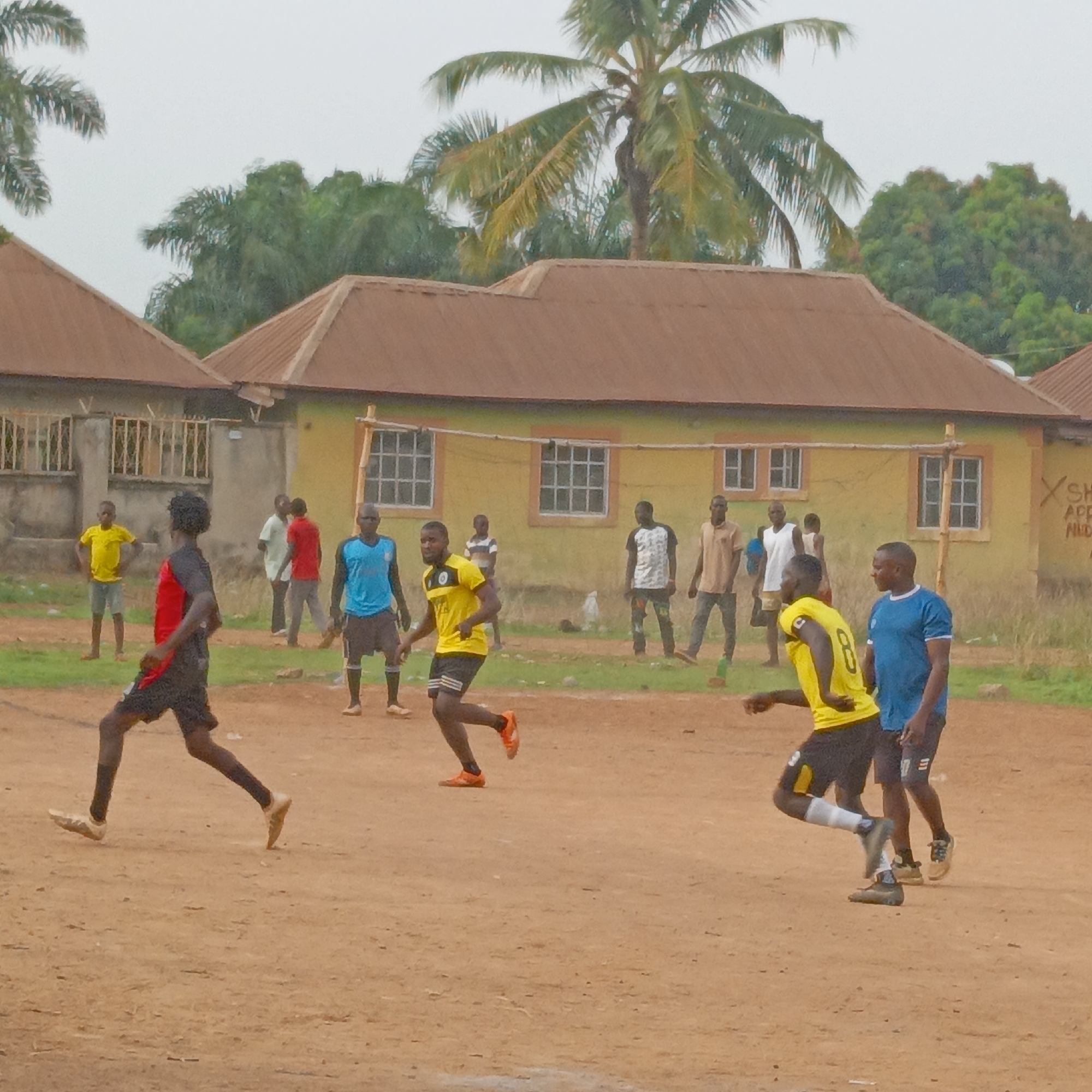 This image captures an informal soccer game underway in Akwanga, Nigeria, under an overcast daytime sky. Approximately 10-12 young men and boys are present, some actively playing on a reddish-brown dirt field, while others stand as spectators or teammates near the background.
In the foreground, a player wearing a black and red kit is shown mid-stride, running from right to left. Further back, another player in a yellow jersey with the number '8' on his back is also in motion. A player in a blue jersey stands near the right side of the field. Several other individuals are scattered in the background, observing the game near the residential structures.
The setting features several simple, single-story houses with corrugated brown roofs. The main building in the background is painted a faded yellow or ochre, featuring multiple white-framed windows and a rudimentary metal gate or fence structure in front. To its left, another building appears to be constructed of grey concrete. The environment is rich with tropical vegetation, including numerous tall palm trees and other green foliage rising above and behind the houses. A makeshift goalpost, constructed from wooden poles, is visible in front of the yellow building.
On the far right wall of the yellow building, some partially visible black text or graffiti can be seen, including "X SH", "APP", and "MID...". The ground is primarily bare earth with sparse patches of green grass along the edges, suggesting a casual, community playing area. The overall scene depicts an active, everyday moment of sport in a local Nigerian setting.