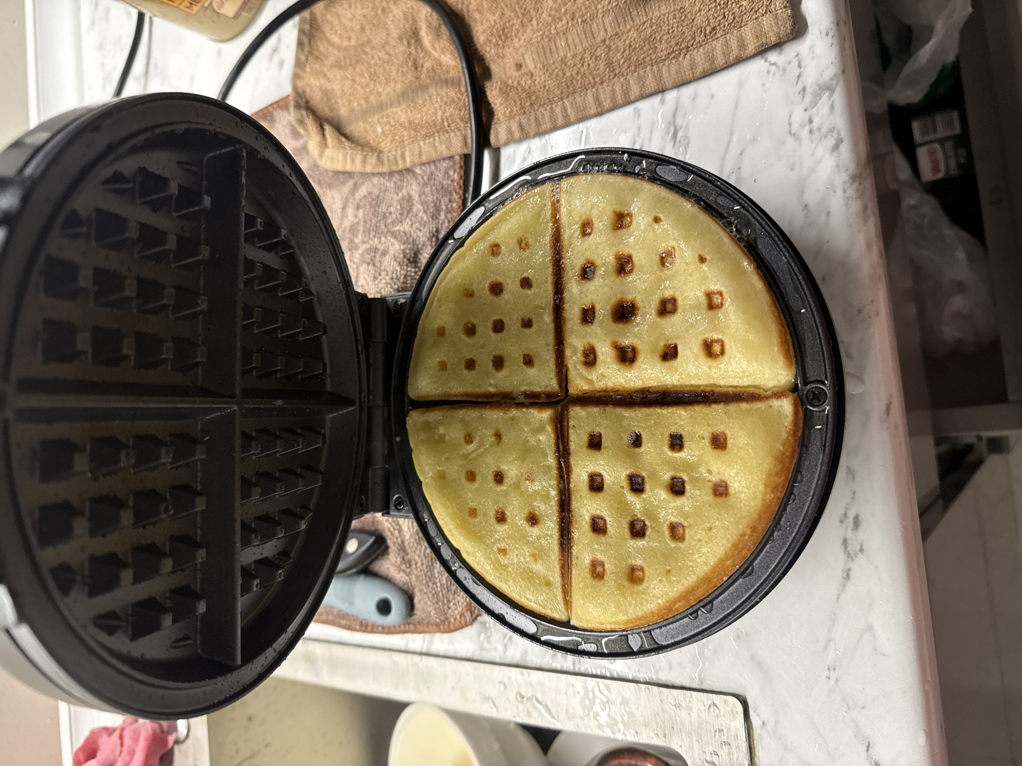 This overhead shot depicts an open, black, circular waffle maker sitting on a white, possibly marble or laminate, kitchen countertop in Batumi, Georgia. The top half of the waffle iron is raised and empty, showing its textured grid pattern, while the bottom half contains a round, freshly cooked waffle.
The waffle is golden yellow and divided into four distinct square sections, with some indentations showing a slightly darker, toasted brown. It appears soft and warm, indicative of recent preparation. The countertop beneath the appliance shows reflections or small drops of liquid, suggesting it might be slightly wet or has just been cleaned.
To the left and slightly behind the waffle maker, a light brown, textured towel is visible. A blue and grey kitchen utensil, possibly a can opener or bottle opener, is also partially visible near the towel. In the background on the right, a cluttered area reveals plastic bags and dark containers, one of which has a faint, illegible label. A small portion of a red towel is visible at the bottom left of the frame. The scene captures a domestic moment of food preparation, likely breakfast or brunch.