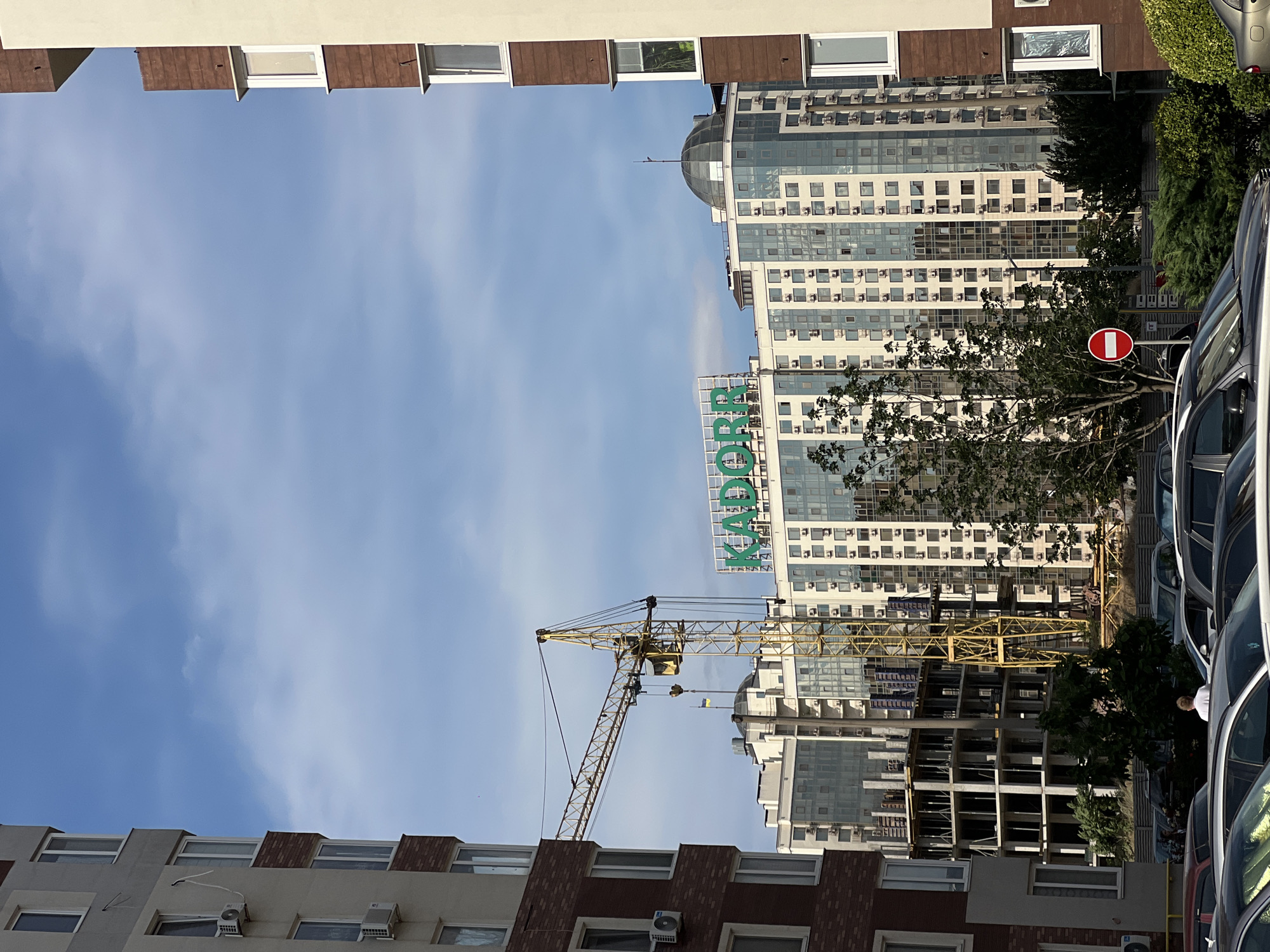 The image captures a street view in Kryzhanivka, Ukraine, featuring a partially completed construction site with a prominent yellow crane. In the background, several modern apartment buildings stand tall, one of which displays a large green sign that reads "KADORR." The sky above is partly cloudy, with patches of blue visible. In the foreground, various cars are parked on the street, suggesting a residential or commercial area. The scene appears to be during the daytime, with natural light illuminating the buildings and surrounding environment.