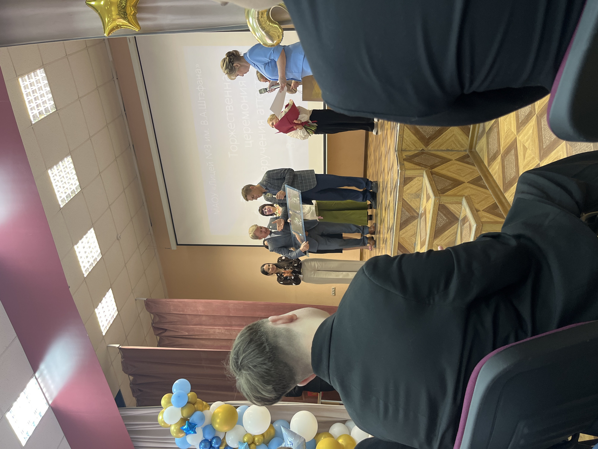 A group of people are gathered on a stage in front of a projection screen. A woman in a light blue uniform is speaking into a microphone and holding a document, appearing to be presenting an award. Another woman in a red jacket stands next to her, holding a bouquet of red flowers. Several people in formal attire are lined up on the stage, some receiving awards or certificates. The projection screen displays text in Russian. The room has a wooden floor and a raised stage. In the foreground, a person wearing a black suit is seated, facing away from the camera. Balloons in blue, gold, and white are arranged near the stage. The lighting suggests an indoor event, likely during the daytime. The scene appears to be an awards ceremony or graduation event. Visible text on the screen includes "МАОУ «Лицей №3 им. В.А. Штифана»" and "Торжественная церемония вручения аттестатов".