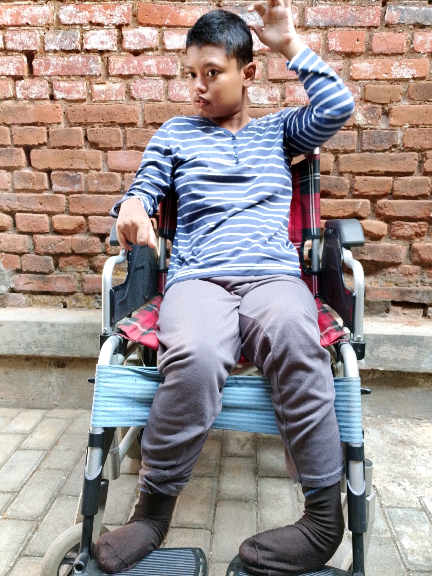 A young person sits in a wheelchair facing the camera, with a brick wall behind them. The person is wearing a blue and white striped long-sleeved shirt and gray pants. Their right arm is raised, and their hand is positioned near their head. The wheelchair has a red and black plaid seat cushion and blue striped straps. The person is wearing dark socks. The ground in front of the wheelchair is paved with light-colored bricks. The location is Banyakan, Indonesia.