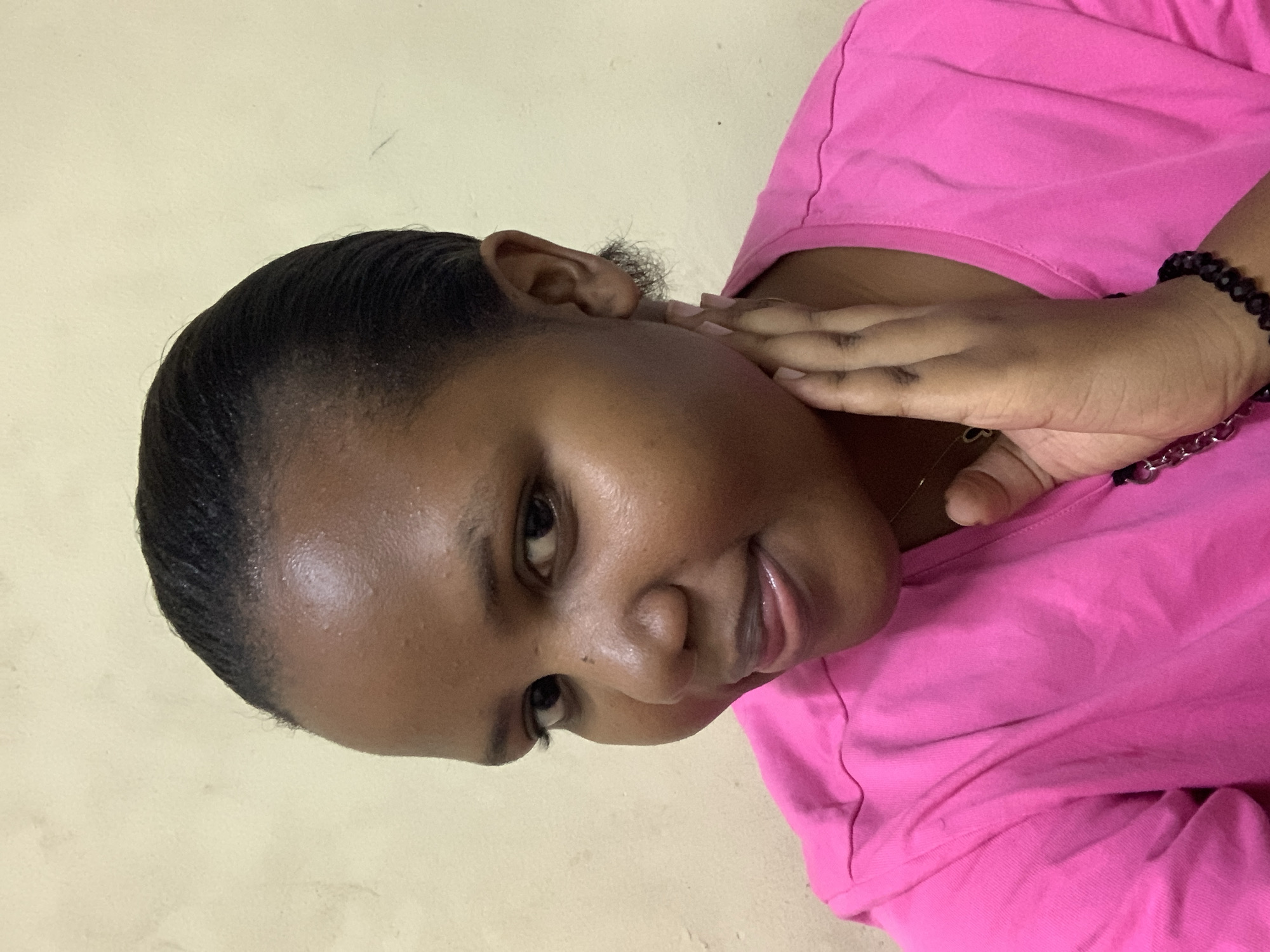 A woman with dark skin is angled towards the viewer, wearing a pink shirt and a black beaded bracelet with a silver chain. She has dark, slicked-back hair and is touching her face with her right hand. The background is a plain, light-colored wall. The location within Dar es-Salaam, Tanzania, is indeterminable.