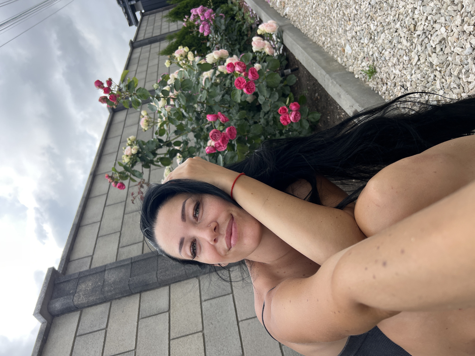 A medium shot, outdoor, selfie-style photograph shows a woman in the foreground, with a rose bush and a grey brick wall in the background. The woman, with long black hair, is looking at the camera with a slight smile. She has on a dark grey strapless top. Her left arm is bent, supporting her head, and her right arm is resting on her lap. A thin red bracelet is visible on her right wrist. The rose bush displays multiple clusters of pink and white roses, with green leaves. The background consists of a grey brick wall with a concrete border, a gravel path, and a cloudy sky. The lighting suggests an overcast day.