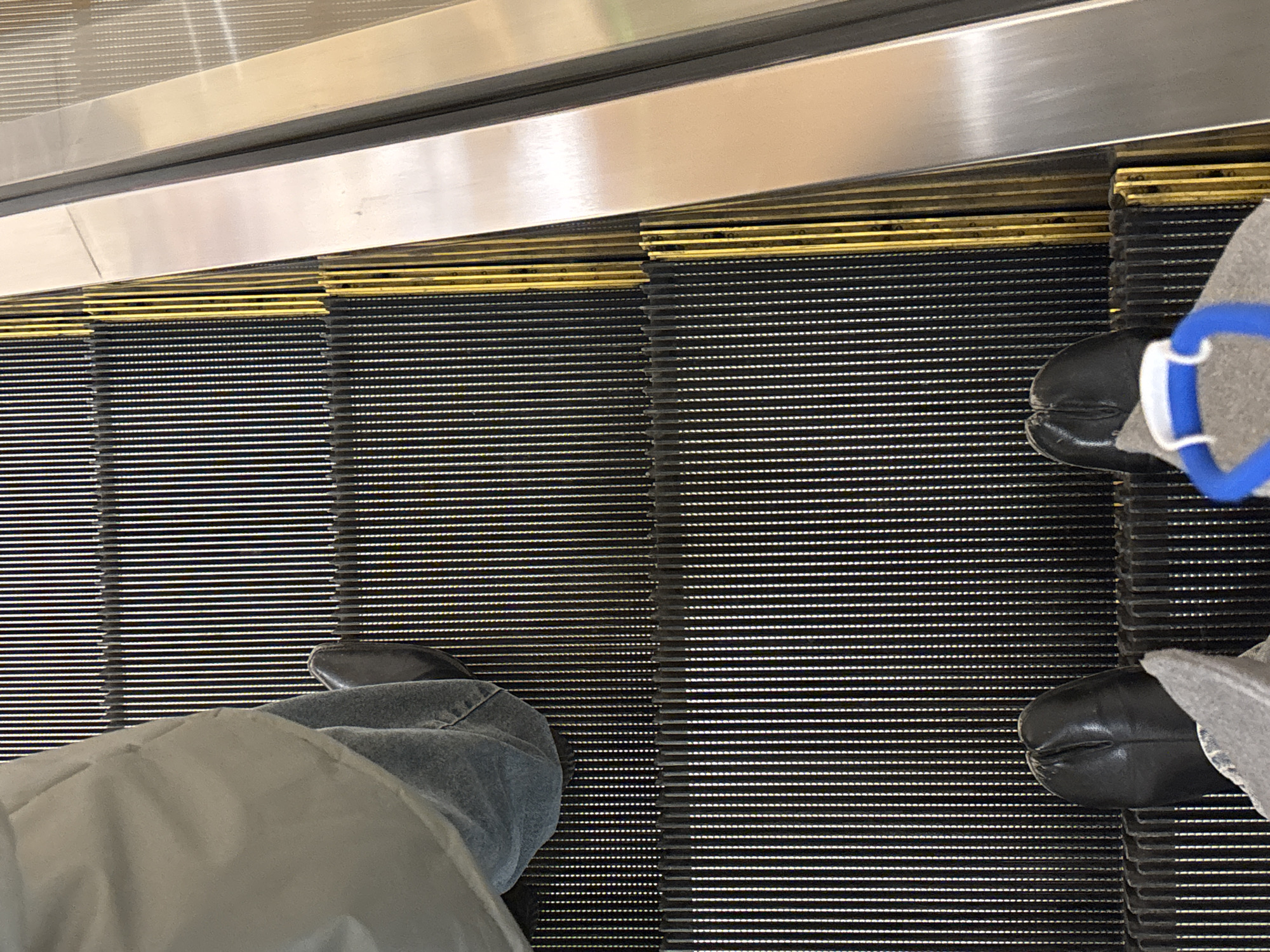 The image depicts a downward-facing view of an escalator in what appears to be a public space. Two individuals are partially visible; one wearing gray jeans and a light gray jacket is positioned on the left, and another wearing gray pants and black shoes with a blue accessory is on the right. The escalator steps are black with yellow edges, and the metallic sides of the escalator are visible at the top of the image. The location is unknown, and there is no definitive evidence to suggest it is in Perm, Russia.
