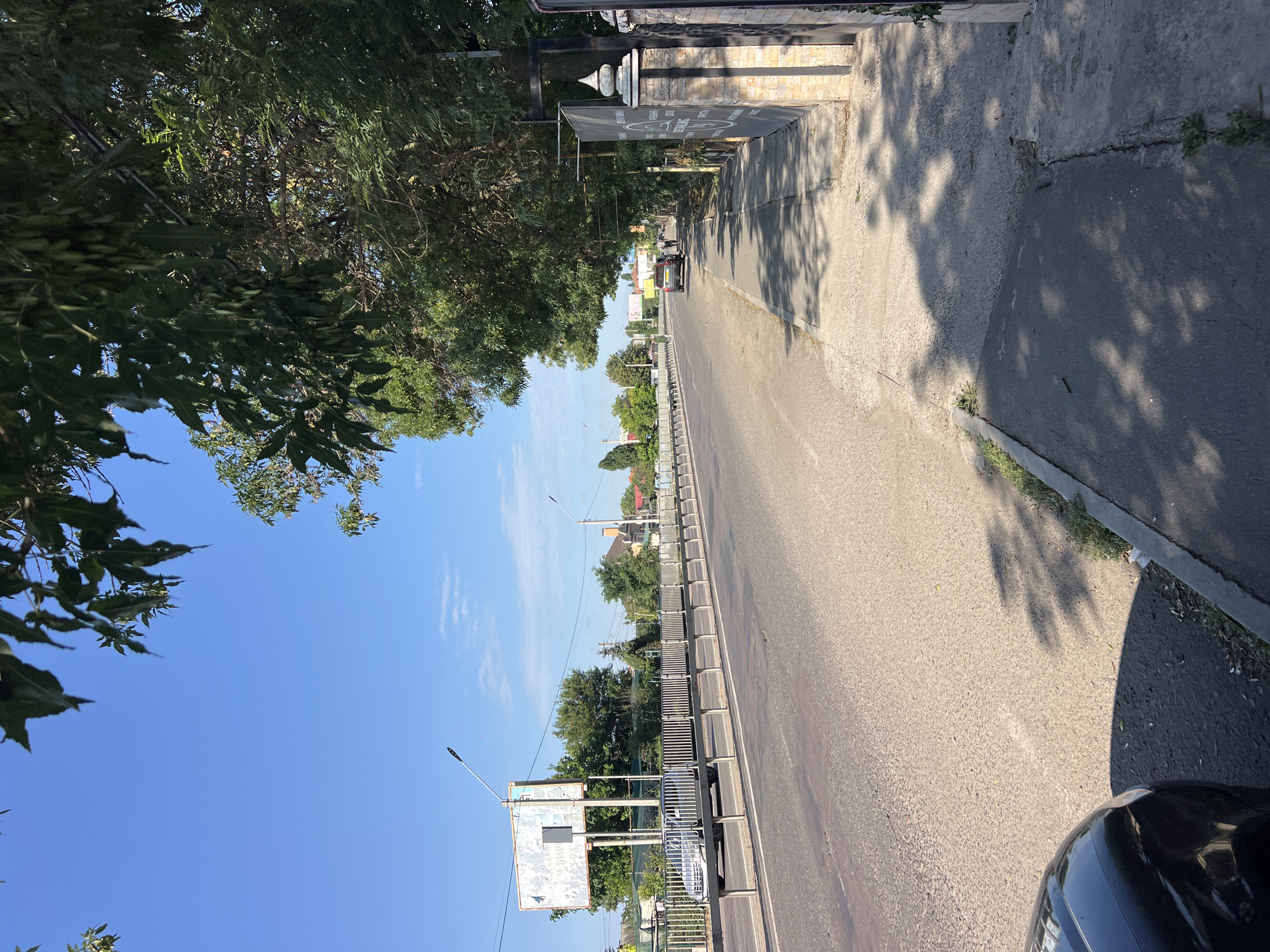 The image shows a street scene in Kryzhanivka, Ukraine, on a clear, sunny day. A paved road runs horizontally across the frame, with a sidewalk to its right. Trees with lush green foliage line the sidewalk and overhang the road, casting dappled shadows on the pavement.
On the road, several vehicles are visible. A silver car is parked on the left side of the road, and further down, a dark-colored car and two individuals are walking along the sidewalk. A white billboard stands on the left side of the road, and a metal railing runs along the side of the road, separating it from a raised embankment. Streetlights are present along the road. The buildings visible in the background appear to be houses and other structures. The sky is a clear blue with a few wispy clouds.