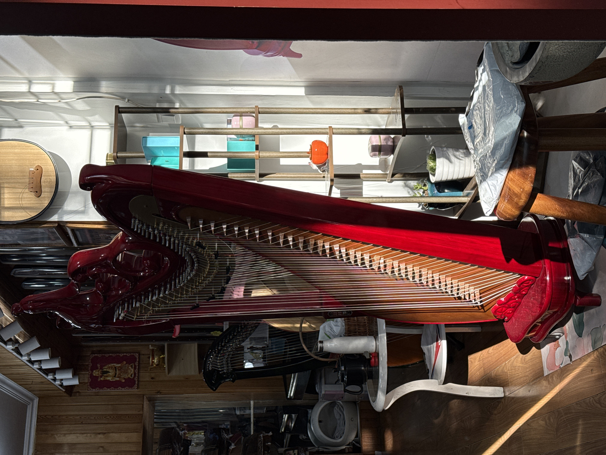A red zither-like instrument is oriented horizontally in a room with wood floors and wood-paneled walls. A second, black zither-like instrument is visible beneath the red one. Various shelves are mounted on the wall containing objects such as a round wooden instrument, planters, and other decorative items. The image is lit by natural sunlight. No people are visible. There is no explicit information regarding the location in Xicheng District, China.