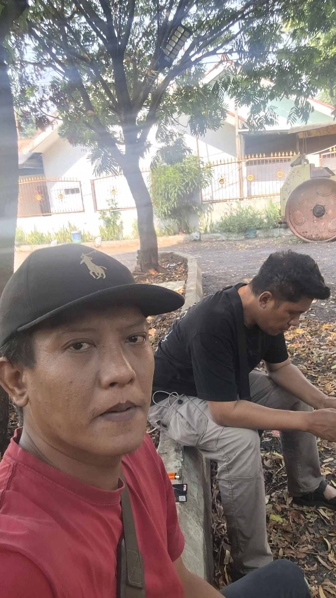 Two individuals are seated outdoors in what appears to be a residential area in Cilaku, Indonesia. The person in the foreground, on the left, is wearing a red t-shirt and a black baseball cap with a gold logo. They are looking directly at the camera. To the right, another individual is seated, wearing a black t-shirt and grey cargo pants, looking down. They are positioned on a concrete ledge, with fallen leaves and dirt on the ground around them. Behind them, a house with a white exterior and a fence can be seen. A large tree with branches spreads over the scene. Part of a large, circular, rusty metal object is visible on the far right. The lighting suggests it is daytime.