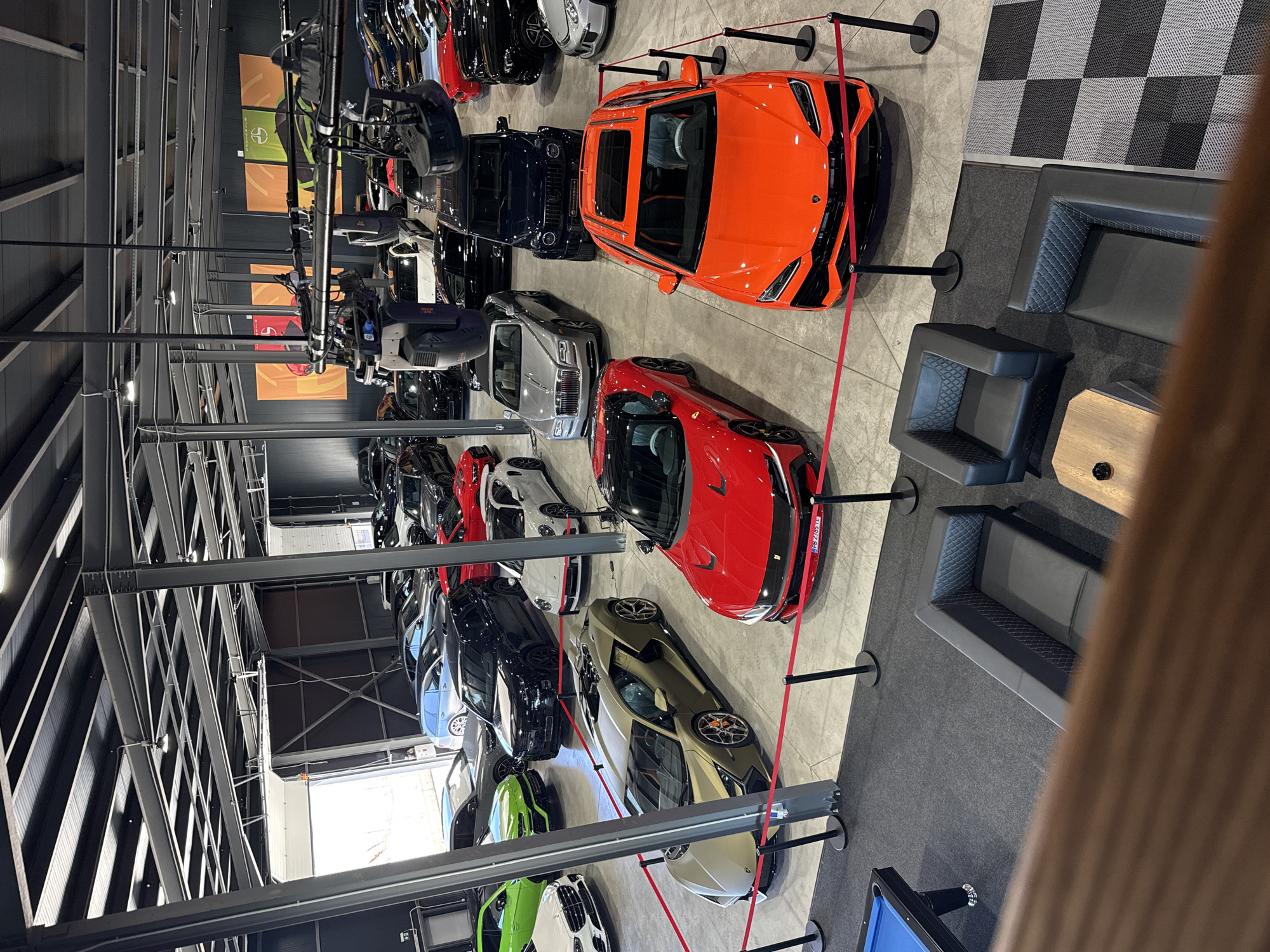 The image depicts a showroom in Plovdiv, Bulgaria, featuring a variety of luxury sports cars. The vehicles, including an orange Lamborghini and a red Ferrari, are arranged in rows and separated by red rope barriers. The cars range in color from bright hues like green and orange to more muted tones of silver, black, and white. The ceiling features a complex grid of metal beams and lighting fixtures. In the foreground, there's a lounge area with dark gray seating and a wooden table. A portion of a blue pool table is also visible. The flooring appears to be gray concrete with a checkered rug section near the seating. The scene is shot from an elevated angle, offering a comprehensive view of the showroom's layout and the cars on display.