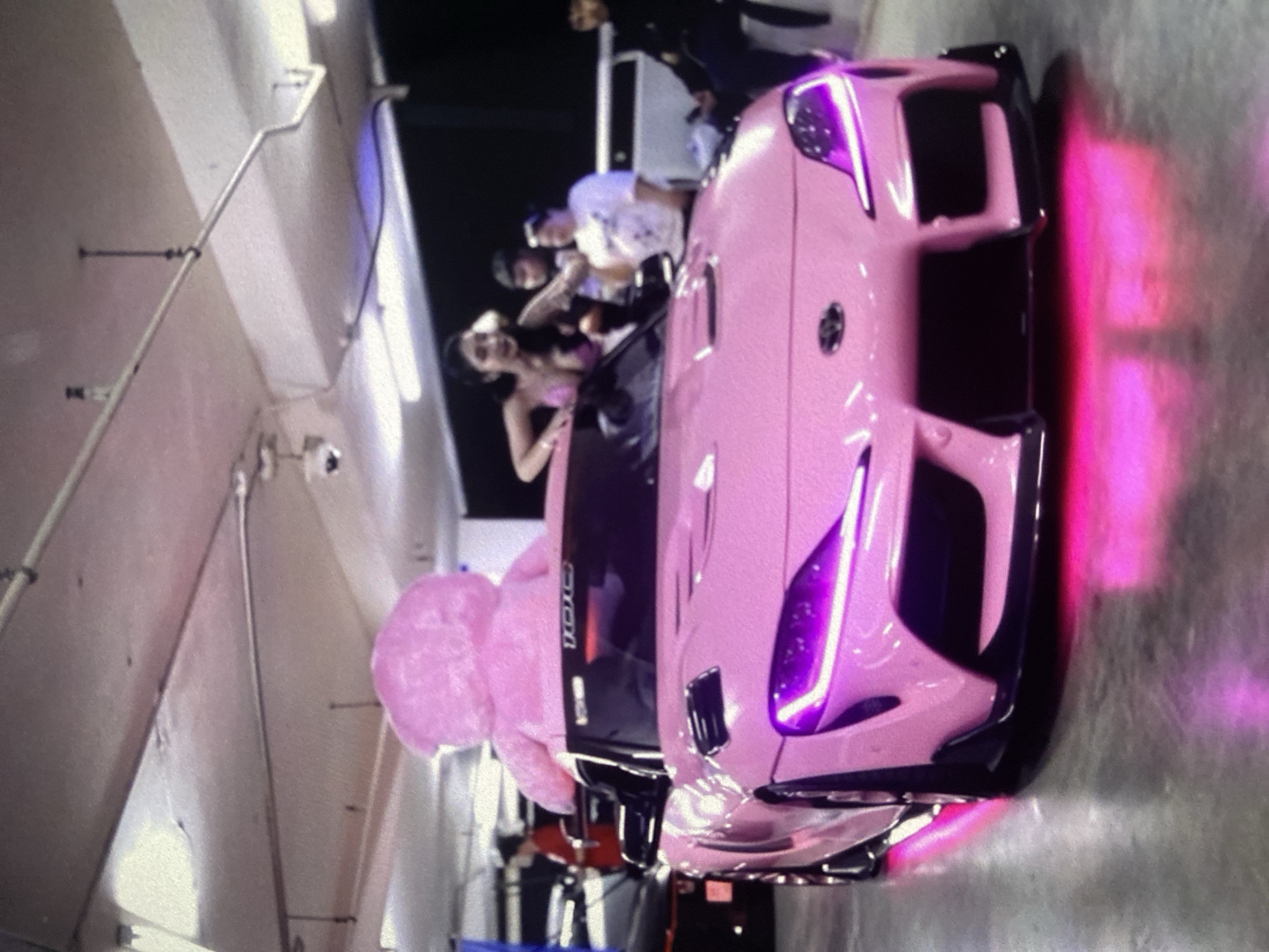 The image shows a pink sports car, possibly a Toyota Supra, parked indoors with several people standing around it. A large pink teddy bear is partially visible near the car's windshield. The car has aftermarket modifications including purple headlights and under-car lighting. The people are looking at or interacting with the car. The indoor location appears to be a parking garage with exposed pipes and lighting fixtures. I cannot confirm its exact location in Lhokseumawe, Indonesia, based on visual information alone.