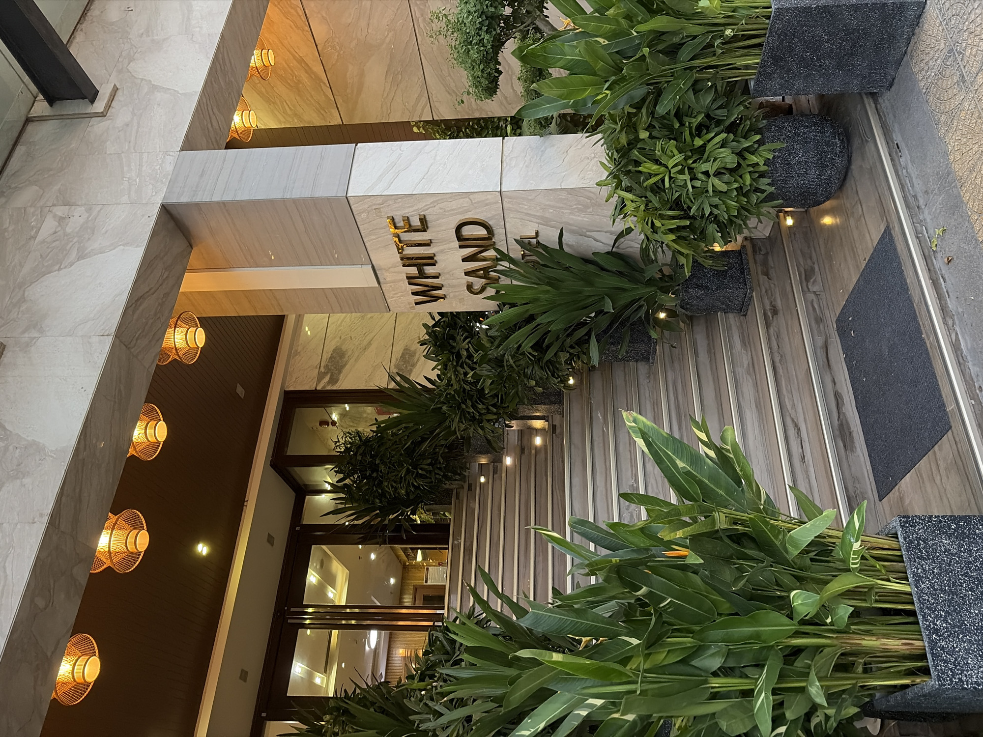 The image shows the entrance to the White Sand Hotel in Đà Nẵng, Vietnam. A set of marble stairs leads up to the hotel's glass doors. Large potted plants with lush green foliage flank the stairs on both sides. Above the entrance, decorative bamboo lamps cast a warm glow. The hotel's name, "WHITE SAND HOTEL," is prominently displayed in gold lettering on a marble pillar. The scene suggests a well-maintained and welcoming hotel entrance.