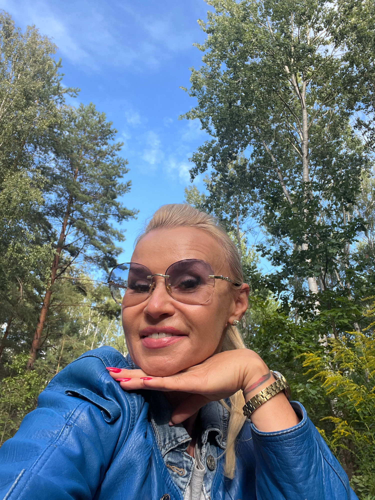 A woman with blonde hair pulled back, wearing large, rimless sunglasses, a blue leather jacket, and a denim shirt underneath, poses for a selfie outdoors. She has her chin resting on her left hand, and her fingernails are painted bright pink. A gold watch is visible on her right wrist. The background consists of tall trees with green leaves and a bright blue sky with scattered white clouds. The scene appears to be in a natural, wooded area, possibly a park or forest. The specific location is Turczyn, Poland.