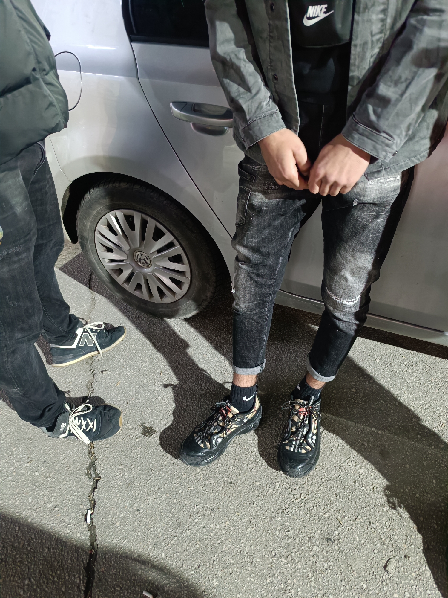 The image shows two individuals standing near a silver Volkswagen car on a paved surface. One individual is wearing black jeans and black New Balance shoes with white laces and "N" logo. The second individual is wearing distressed black jeans, black Nike socks, and black Burberry sneakers with patterned laces. A black Nike bag is visible under the gray jacket worn by this person. The setting appears to be an urban environment, potentially in Asenovgrad, Bulgaria. A crack is visible on the paved surface next to one of the pairs of sneakers. The overall lighting suggests daytime conditions.