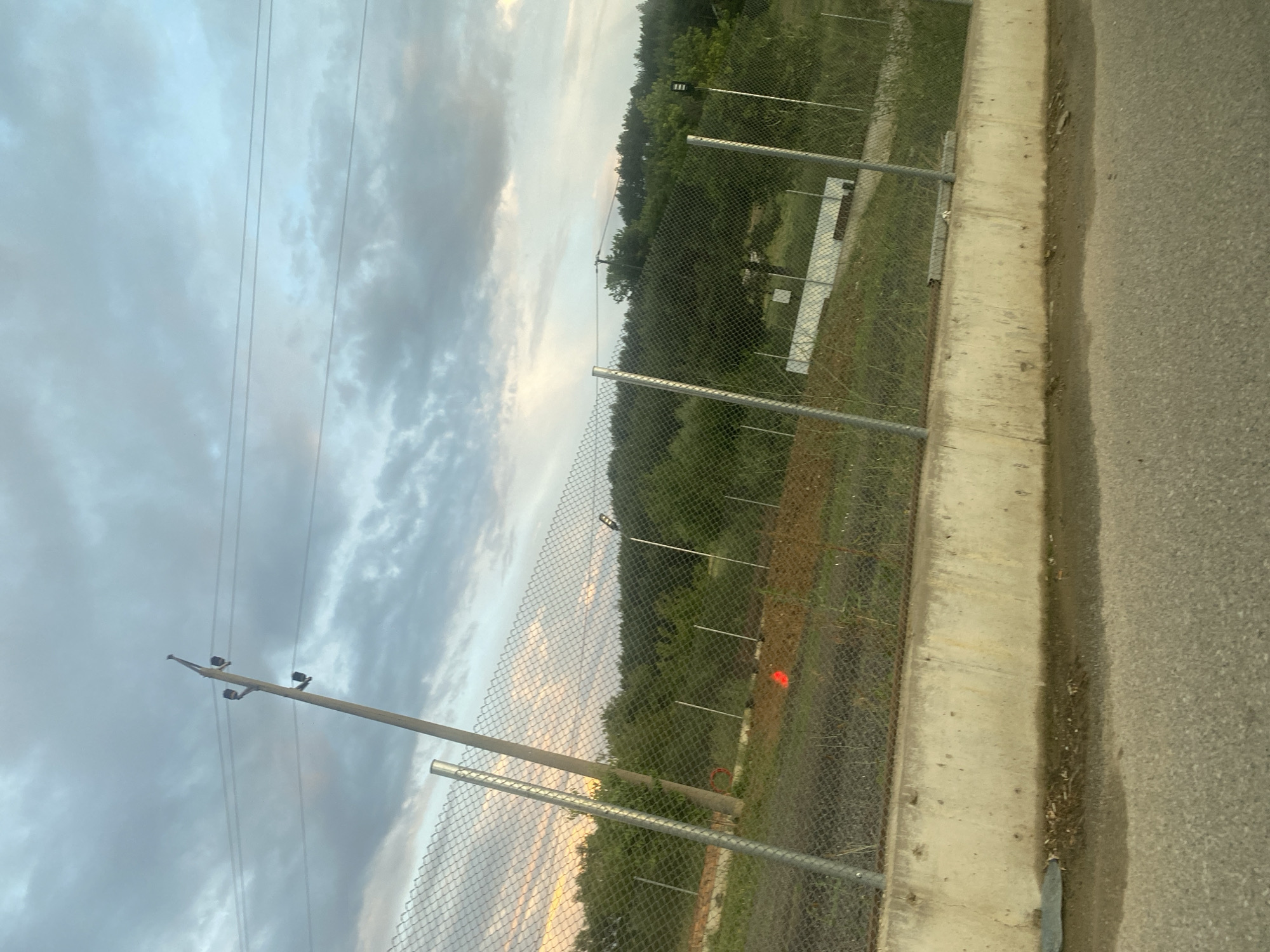 The image depicts an outdoor scene, likely in the vicinity of Ablanitsa, Bulgaria, under a cloudy sky. In the foreground, a paved surface, possibly a road or pathway, is visible, bordered by a concrete barrier. Beyond the barrier, a chain-link fence runs horizontally across the frame, separating it from the background. The fence is supported by metal posts.
Behind the fence, the terrain slopes upwards, covered in grass and dirt. Scattered throughout this area are various objects including what appear to be construction materials or debris, and some red markers or flags. A cluster of trees and dense foliage forms a natural backdrop, with a more distant line of trees suggesting a forest or heavily wooded area. Two prominent utility poles, one leaning significantly, are visible against the sky, with wires extending between them. One of the utility poles has what appear to be insulators attached. Further in the distance, a white structure or building can be discerned amidst the trees, and a light fixture is mounted on a pole. The overall lighting suggests it is either early morning or late evening, given the soft illumination and the color of the sky. There are no people or animals clearly visible in the image.