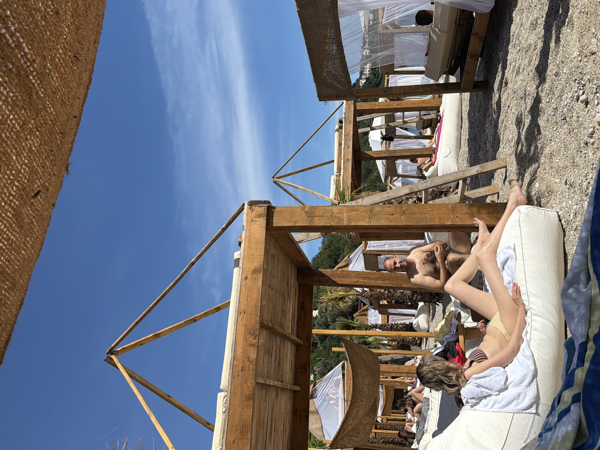 The image captures a beach scene, likely taken in Drobnići, Montenegro, judging by the landscape and architectural elements. The scene is dominated by a series of wooden beach cabanas, constructed with exposed wooden beams and some draped with sheer white fabric and woven sunshades.
In the foreground, a man and woman are reclining on a white lounge chair. The man is shirtless and faces the viewer. The woman is lying down as well. Other figures are visible in the background, also lounging in cabanas.
The environment is a mix of beach and vegetation. The ground is covered with pebbles. In the distance, lush green vegetation and hills are visible. The sky is a clear blue with wispy clouds.