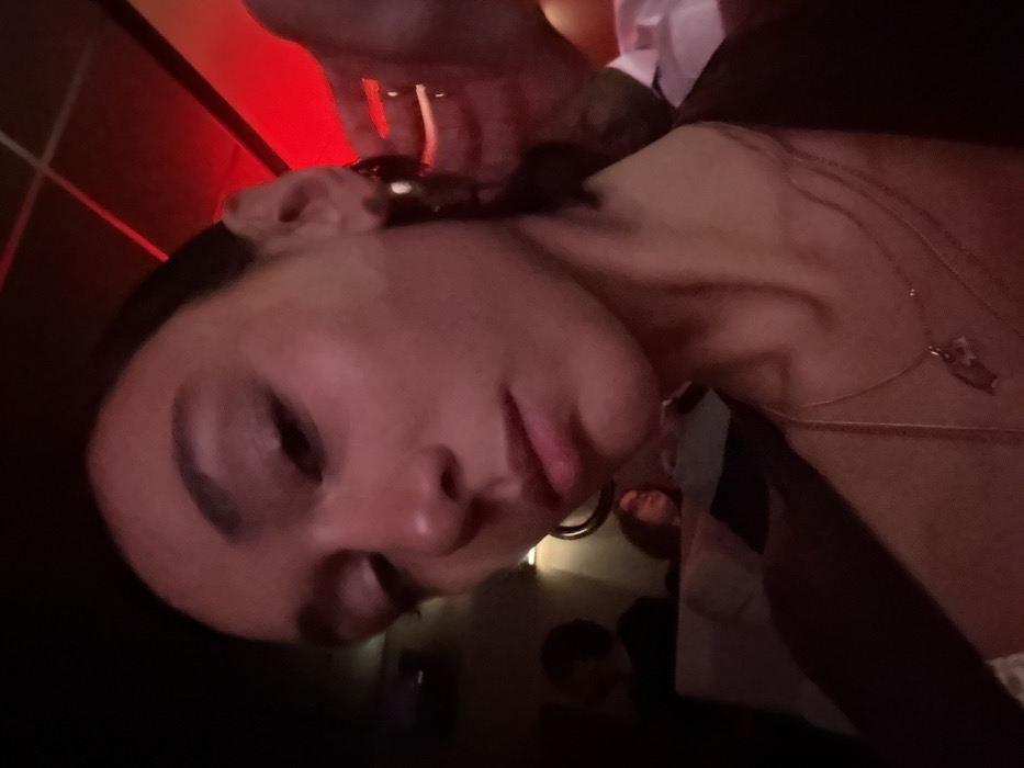 A woman's face dominates the frame, illuminated by red lighting from above and possibly behind. Her expression is subtle, and her dark hair is pulled back. A hand with rings on the fingers is visible near her head. She wears gold necklaces, and a glimpse of another person is seen in the background. The overall composition has a dark, intimate, and slightly blurred quality.