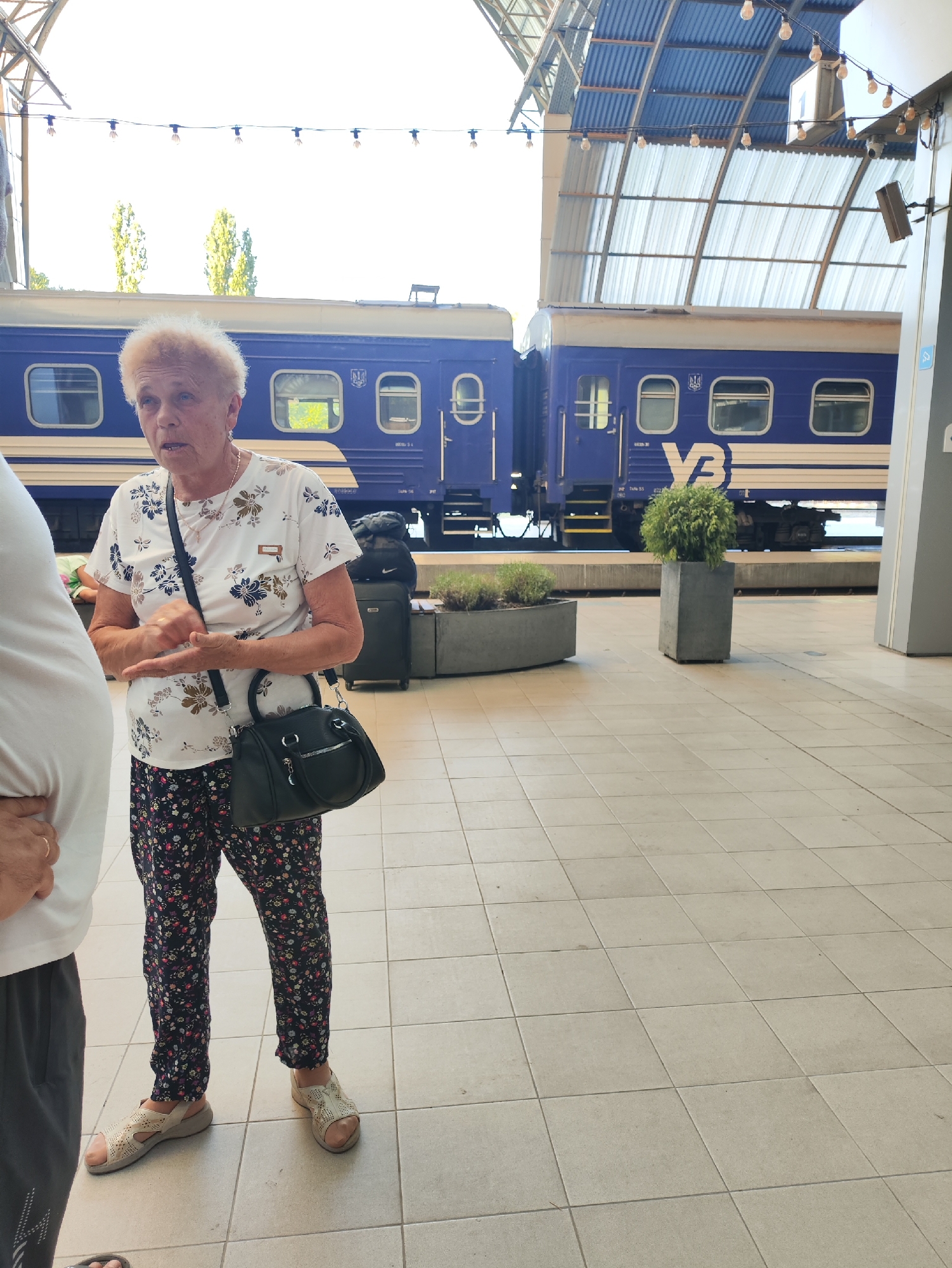 A medium shot captures an older woman standing on a tiled platform in what appears to be a train station. She is wearing a white floral-patterned t-shirt and dark floral-patterned pants, accessorized with beige sandals and a black handbag slung over her shoulder. Her hair is styled in a voluminous blonde perm, and she is looking slightly to her left, her mouth open as if speaking. Behind her, a blue train with white stripes and windows stretches along the platform, partially obscured by the station's roof structure, which features corrugated metal roofing supported by a metal framework. The environment suggests an outdoor covered area, with exposed light bulbs strung across the ceiling. The presence of potted plants, one a round green shrub in a concrete planter, adds a touch of greenery to the scene. The overall impression is a candid moment at a transit hub, possibly during daylight hours due to the bright illumination. The location is not specified as Chișinău, Moldova.