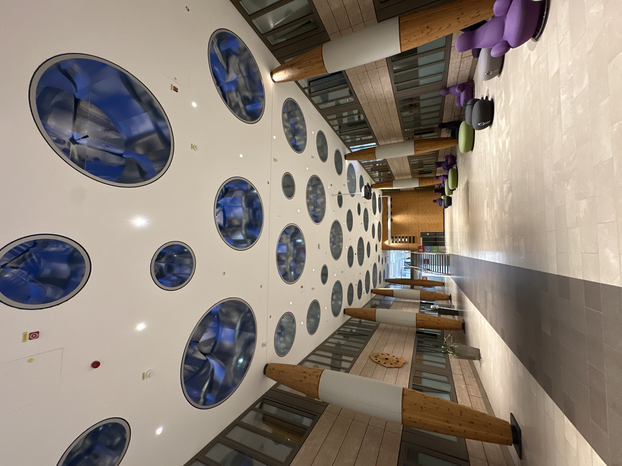 The image shows a modern, spacious interior hallway. The ceiling is white with numerous large, oval-shaped recessed lights that reflect a blue and silver pattern. Along the sides of the hallway are large, slanted wooden pillars with white cylindrical bases. The walls are clad in light brown wood-paneled material and feature rows of windows. The floor is tiled with large, light-colored rectangular tiles. In the distance, a staircase is visible, along with some modern, colorful seating arrangements, including purple chairs and green and grey beanbag-like stools. A few plants in grey planters are also present. The lighting is bright and diffused, creating a clean and contemporary atmosphere. There are no people visible in the immediate foreground or midground. The scene is indoors, likely in a public building such as an office, educational institution, or commercial space. The image does not contain any visual cues to indicate it is located in Garmisch-Partenkirchen, Germany.