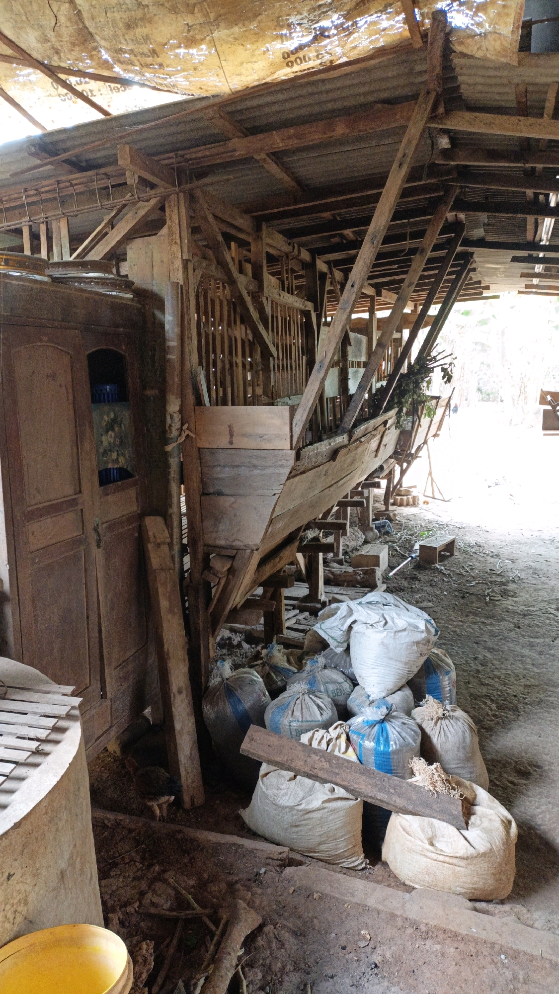 The image depicts an interior scene of a wooden structure, possibly a barn or workshop, in Suwawal Timur, Indonesia. The structure features a rudimentary roof made of corrugated metal sheets and what appears to be a tarp or plastic sheeting, supported by wooden beams. A wooden staircase or elevated platform is visible, constructed with rough-hewn planks and posts, suggesting a functional but unrefined building style.
In the foreground, a pile of large, white and grey sacks is stacked on the ground. Some of these sacks have blue banding. A rough wooden plank rests horizontally across the top of several sacks, and some loose wood shavings or debris are visible on and around the sacks.
To the left of the sacks, a tall, wooden cabinet or wardrobe with two doors stands against a wooden post. The cabinet is weathered and appears to be made of dark wood.
The floor of the structure is covered in dirt and debris, with scattered pieces of wood and what looks like dried leaves. In the lower left corner, a portion of a large, yellow, cylindrical object, possibly a bucket or container, is visible.
Further into the structure, more wooden supports and beams are visible, as well as what looks like the beginnings of a raised enclosure or pen, possibly for livestock, with vertical wooden slats. A bird, likely a chicken or fowl, is standing on the ground between the sacks and the cabinet.
The background shows more of the wooden structure and a bright, sunlit area beyond, suggesting an opening or the exterior of the building. The overall impression is one of a rural or agricultural setting, with evidence of manual labor and storage of materials.