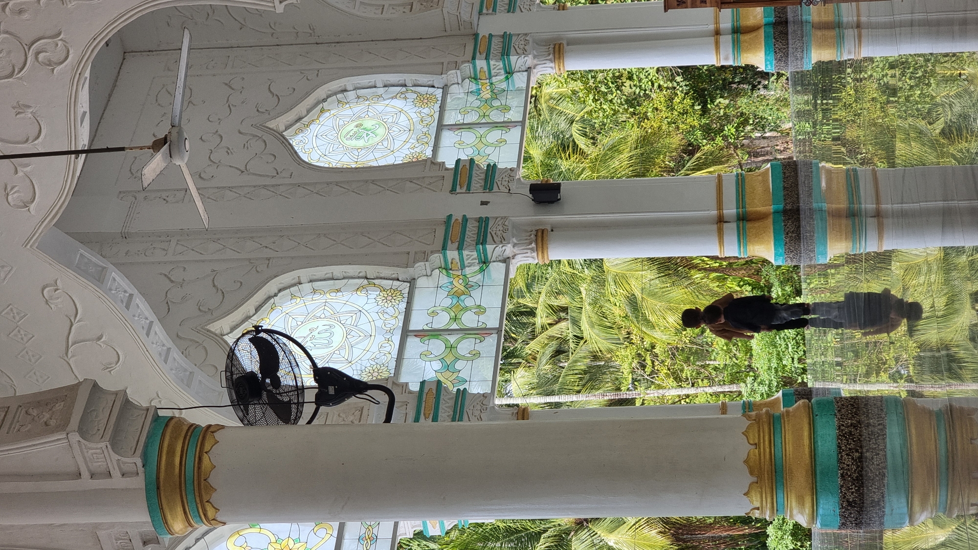 Two young boys walk away from the camera towards a lush green tropical landscape visible through ornate windows and archways of a building, likely a mosque, located in Lapang Timu, Indonesia. The building features white pillars with gold and teal decorative bands, intricate white plasterwork on the ceiling, and stained-glass windows with Islamic calligraphy. A black oscillating fan is mounted on a pillar, and a ceiling fan is visible in the background. The floor is highly reflective, mirroring the pillars, ceiling, and the scene outside, creating a shimmering effect. The boys appear to be dressed casually, one in a brown shirt and the other in a black shirt.
