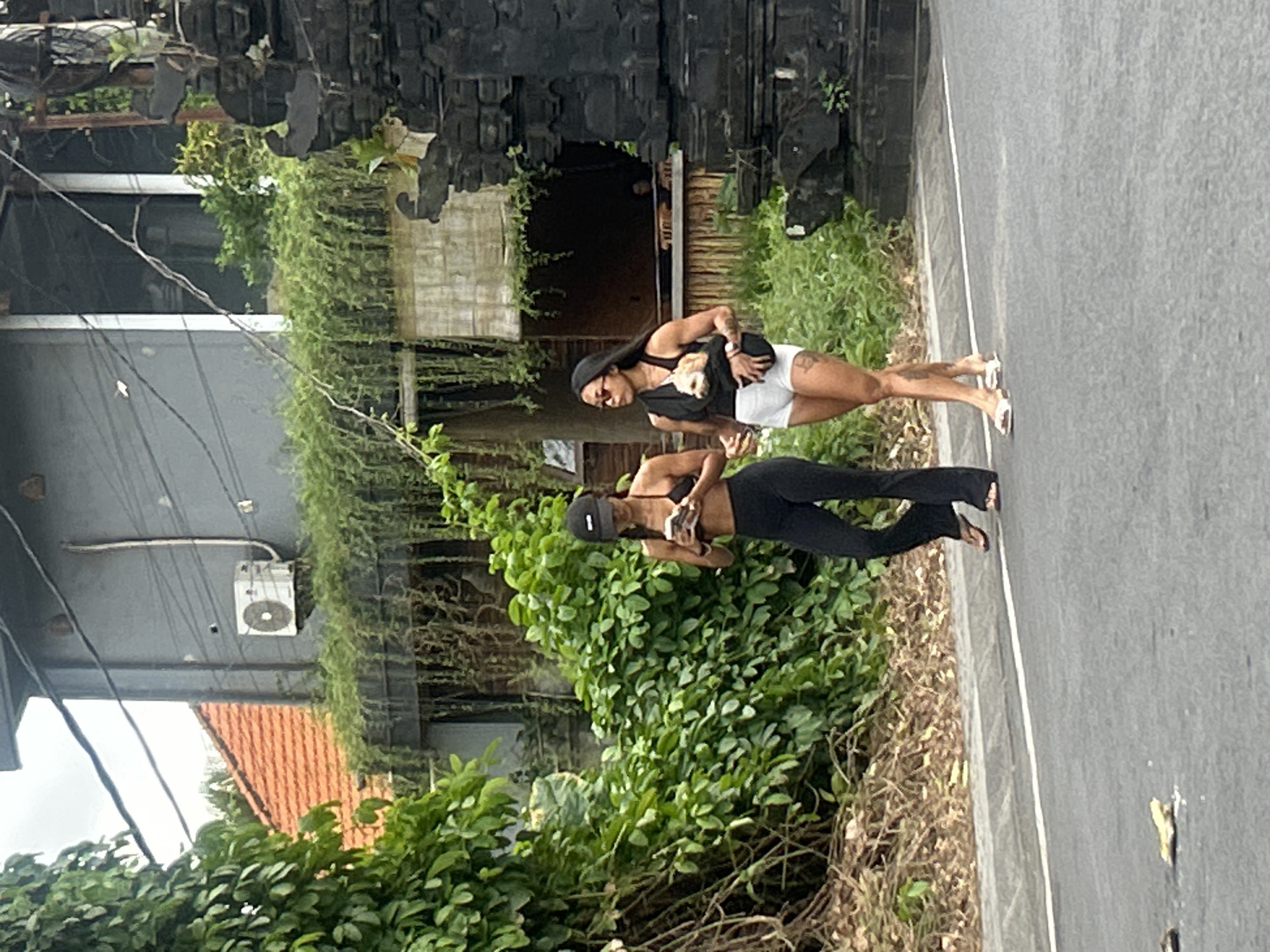 Two women are walking on a paved road in a tropical setting. The woman in the foreground, wearing a black baseball cap, black crop top, and black flared pants, is carrying a phone in her hand. The second woman, behind her, is wearing a black tank top, white shorts, and a black fanny pack slung across her chest. She has a small dog in her arms. The scene is lush and green, with dense foliage bordering the road. In the background, there are hints of traditional Indonesian architecture with dark, carved stone structures. The sky appears overcast, suggesting it is daytime. The overall mood seems casual and relaxed.