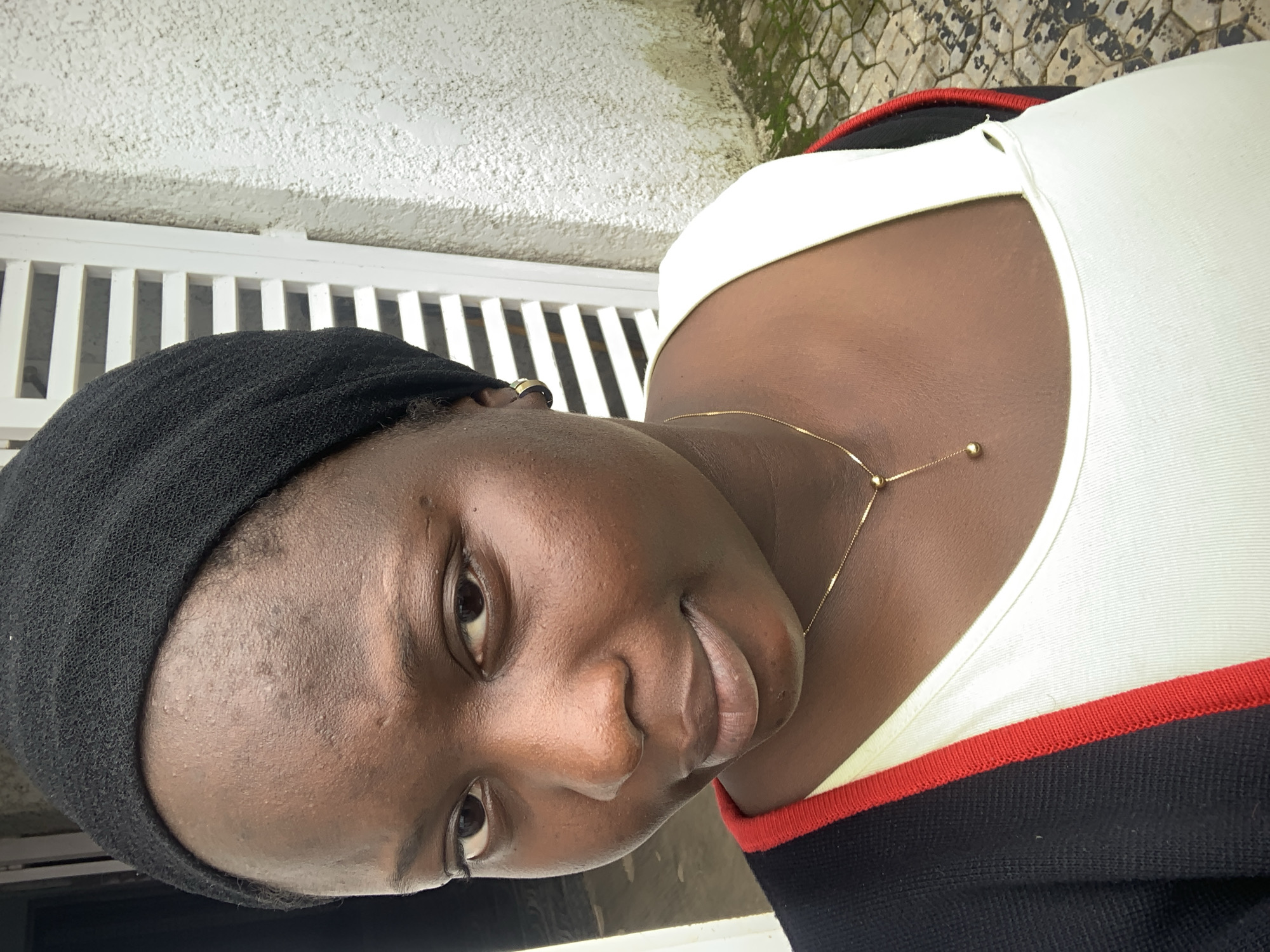 An outdoor, close-up, selfie-style photograph captures a young Black woman from the chest up. She is wearing a black headscarf, a white tank top, and a black cardigan with red trim. A delicate gold necklace with a small bead pendant rests on her collarbone. Her skin is a rich, dark brown, and she has dark brown eyes and dark hair peeking out from beneath the headscarf. She is looking directly at the camera with a slight smile. The background consists of a textured, off-white wall and a white slatted structure, possibly a fence or shutter, on the left. Paving stones are visible on the ground at the bottom right of the frame. The lighting suggests it is daytime.