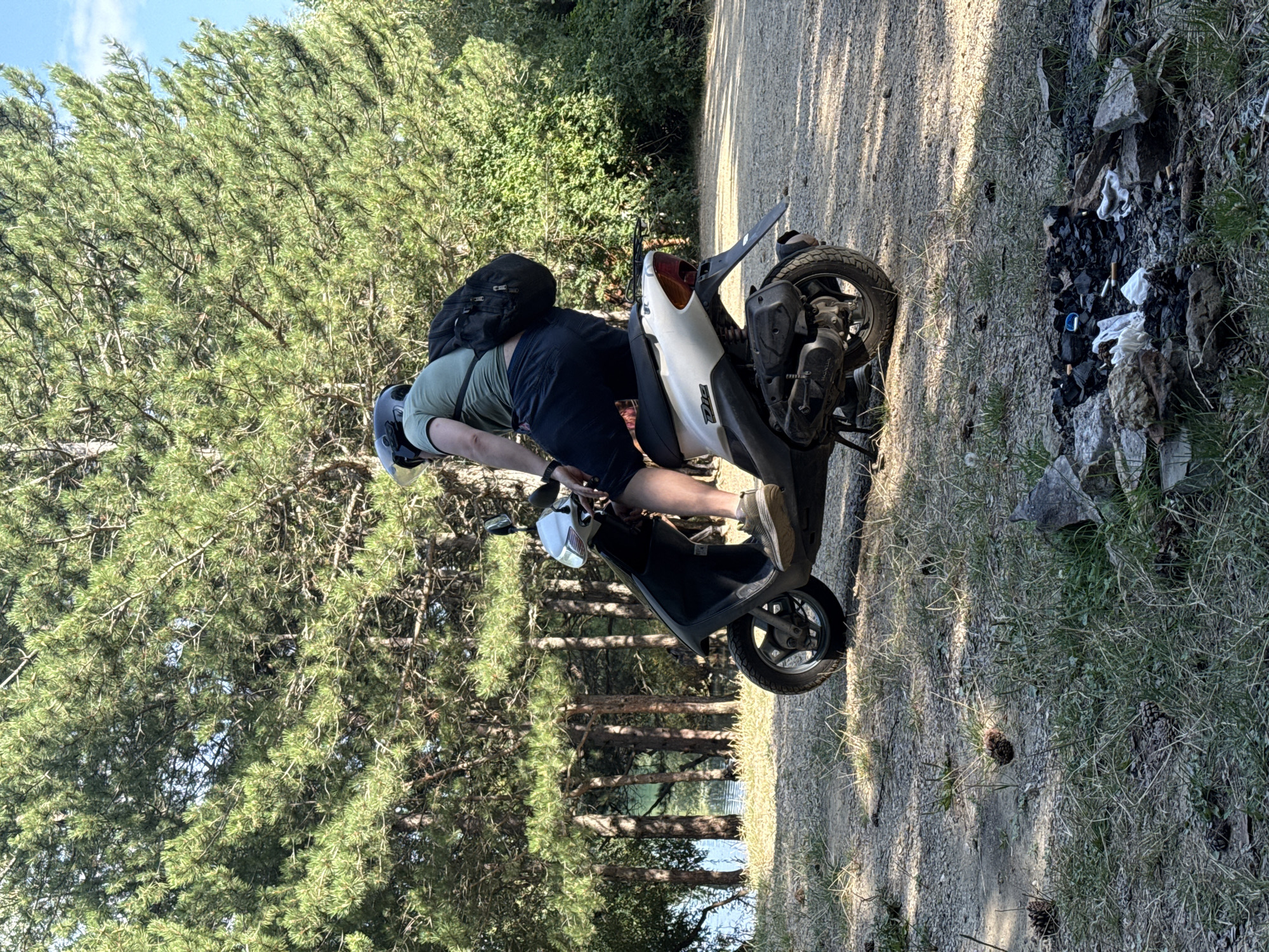 A person wearing a helmet and a backpack is seated on a scooter. The scooter is parked on a dirt path next to a body of water. Tall pine trees and green bushes surround the area. In the foreground, there is a fire pit with remnants of a fire and scattered cigarette butts. The image appears to have been taken in Zaprudnoe, Ukraine, on a sunny day.