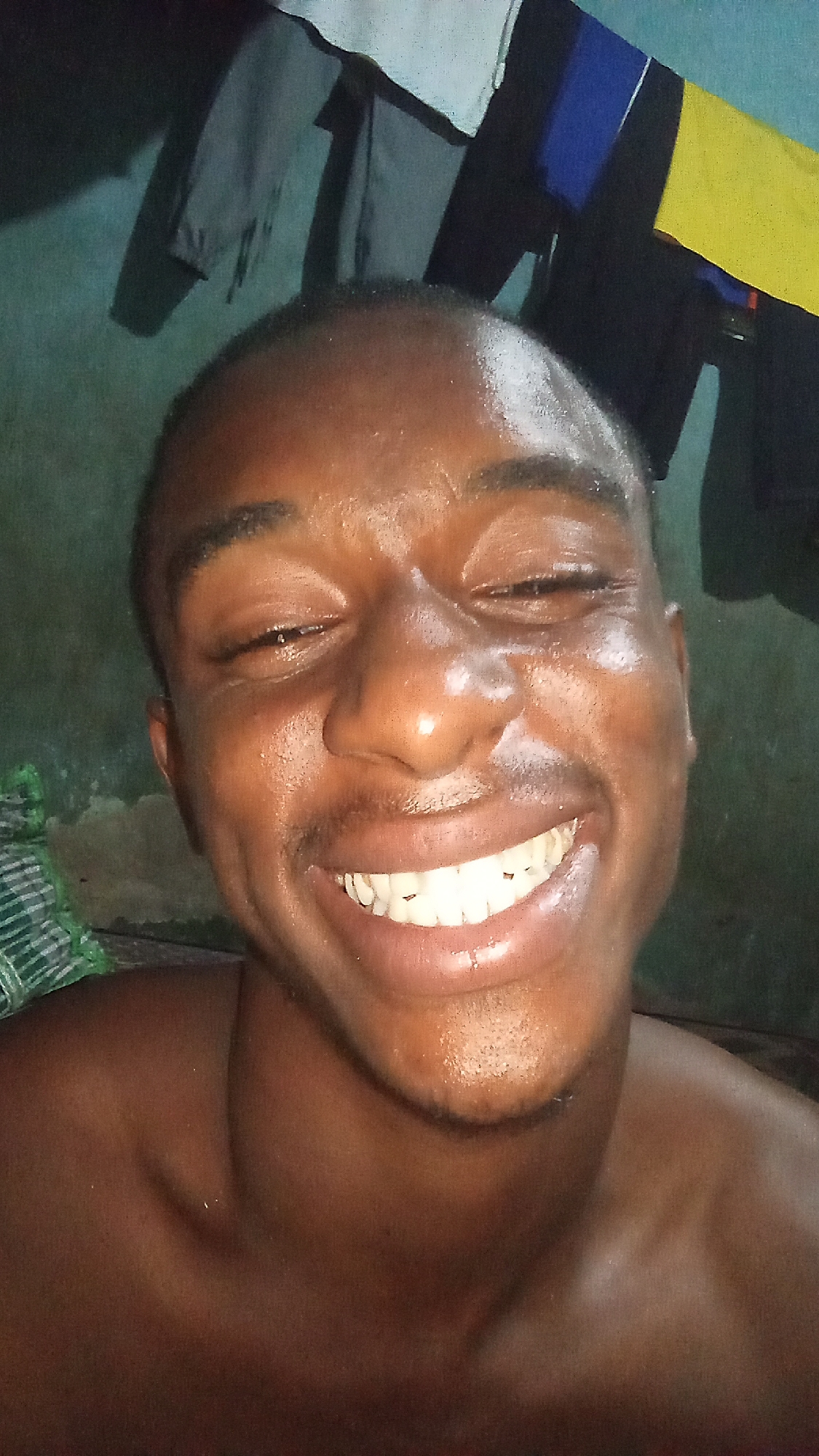 A close-up, head-and-shoulders shot captures a smiling young Black man. The man is facing the camera with a slight upward tilt. He has dark skin, close-cropped hair, and is shirtless. The man's face appears damp. Behind the man, out of focus, are clothes hanging to dry. The environment suggests a residential setting. There is no information that indicates that this image was taken in Onitsha, Nigeria.