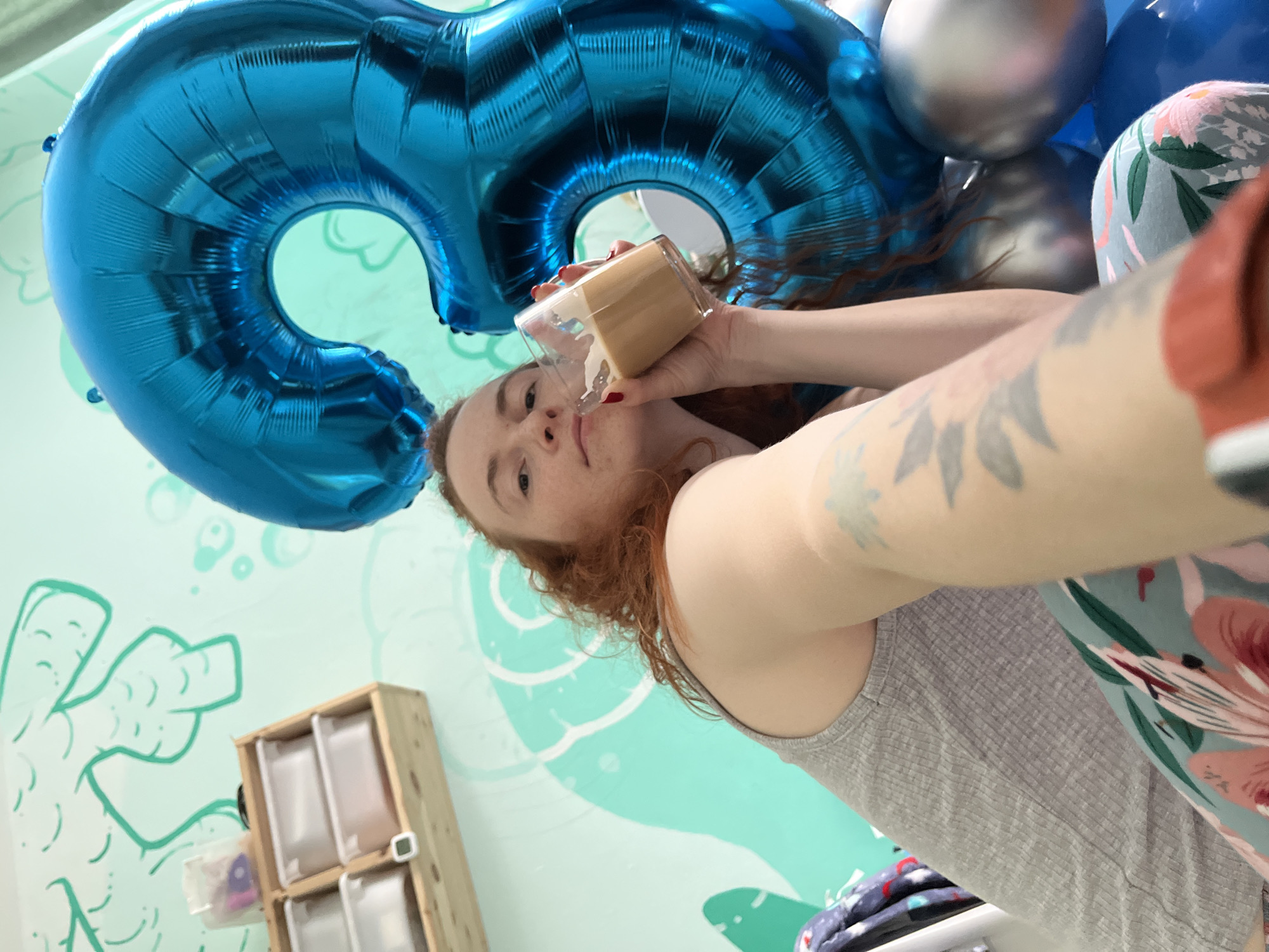 A selfie of a pale-skinned woman with red hair and freckles, wearing a gray tank top and floral pants, taken indoors. She is holding a clear glass partially filled with a tan liquid, possibly a beverage, near her mouth. Behind her, a large blue balloon in the shape of the number "6" is visible, along with other balloons. The background shows a light green wall with a cartoonish drawing. A wooden storage unit with items inside can be seen in the lower-left corner. The location is likely Chaikovsky, Russia.