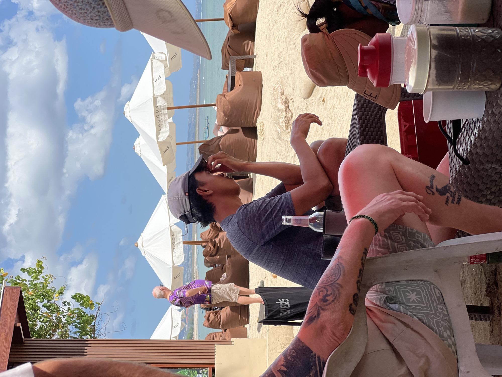 A group of people are seated at a beach lounge in Kuta, Indonesia, under a bright, sunny sky with scattered clouds. The beach is sandy, with several large, white umbrellas providing shade. Beanbag chairs are arranged on the sand.
In the foreground, a person with extensive tattoos on their arm and leg is seated on a white plastic chair. They are wearing light-colored shorts with a patterned fabric underneath. Another person, wearing a dark grey t-shirt and a grey baseball cap, is seated to the right, facing away from the camera and appearing to be touching their face. A bottle is placed in a holder on the table in front of them.
In the middle ground, a man wearing a vibrant purple t-shirt and light-colored shorts stands looking towards the ocean. The "Lagoon Lounge" logo is visible on one of the umbrellas.
In the background, the turquoise ocean stretches out, with a few boats visible on the water. The coastline is visible in the distance. On a table in the foreground, there are several containers, including a red-lidded shaker bottle and a clear jar filled with dark material, alongside a white cup. The overall atmosphere is relaxed and suggests a beachside resort setting.