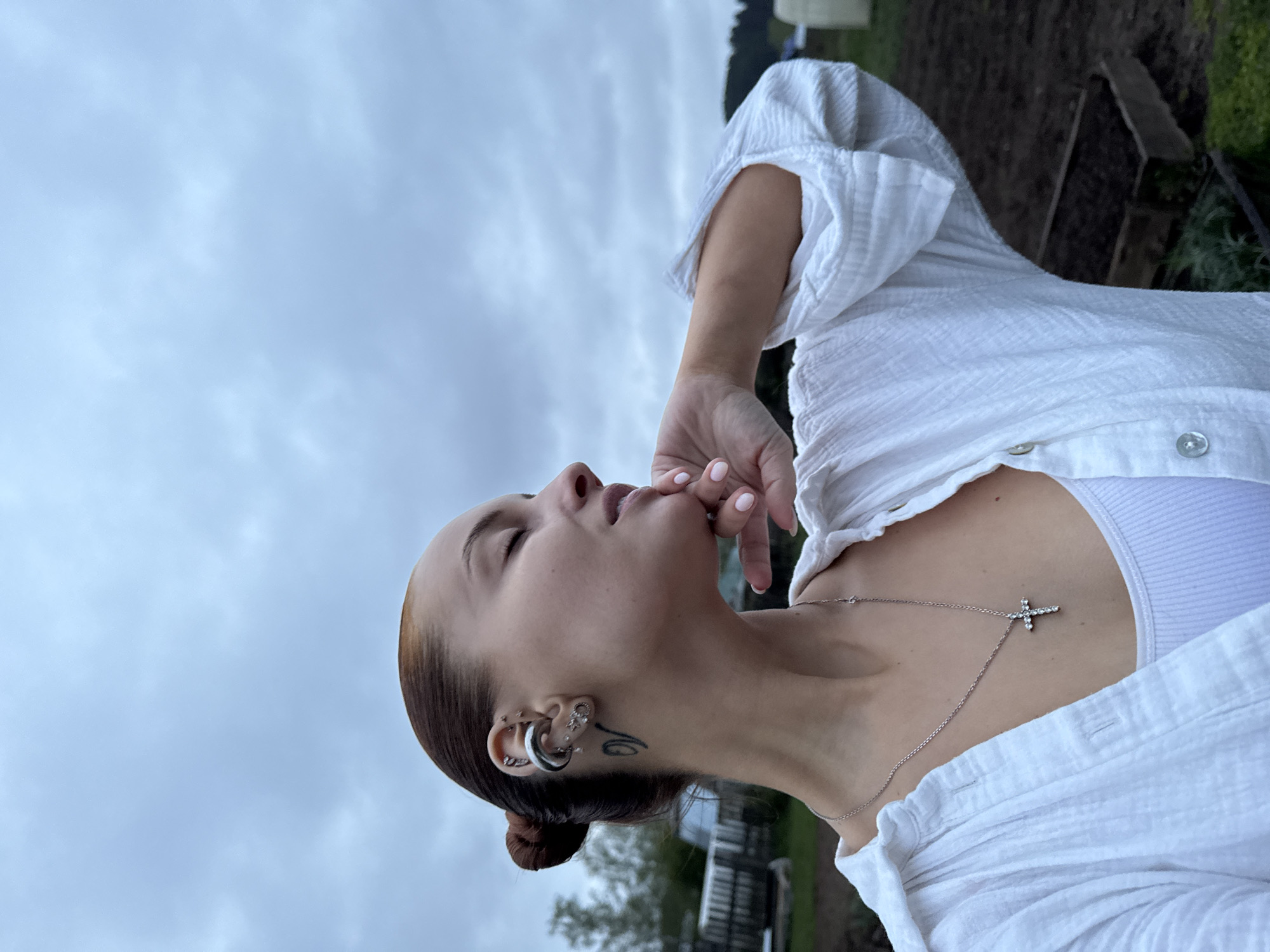 A young woman is depicted outdoors, likely in a rural or suburban area of Chastyye, Russia, judging by the visible vegetation and sky. She is wearing a white, textured button-down shirt over a white tank top. A silver necklace with a cross pendant is visible around her neck. The woman has brown hair styled in a bun, and several ear piercings are present, along with a tattoo on the side of her face. Her right hand is raised to her mouth, with her fingers touching her lips. The background includes a cloudy sky, green grass, and what appears to be a garden bed or field.