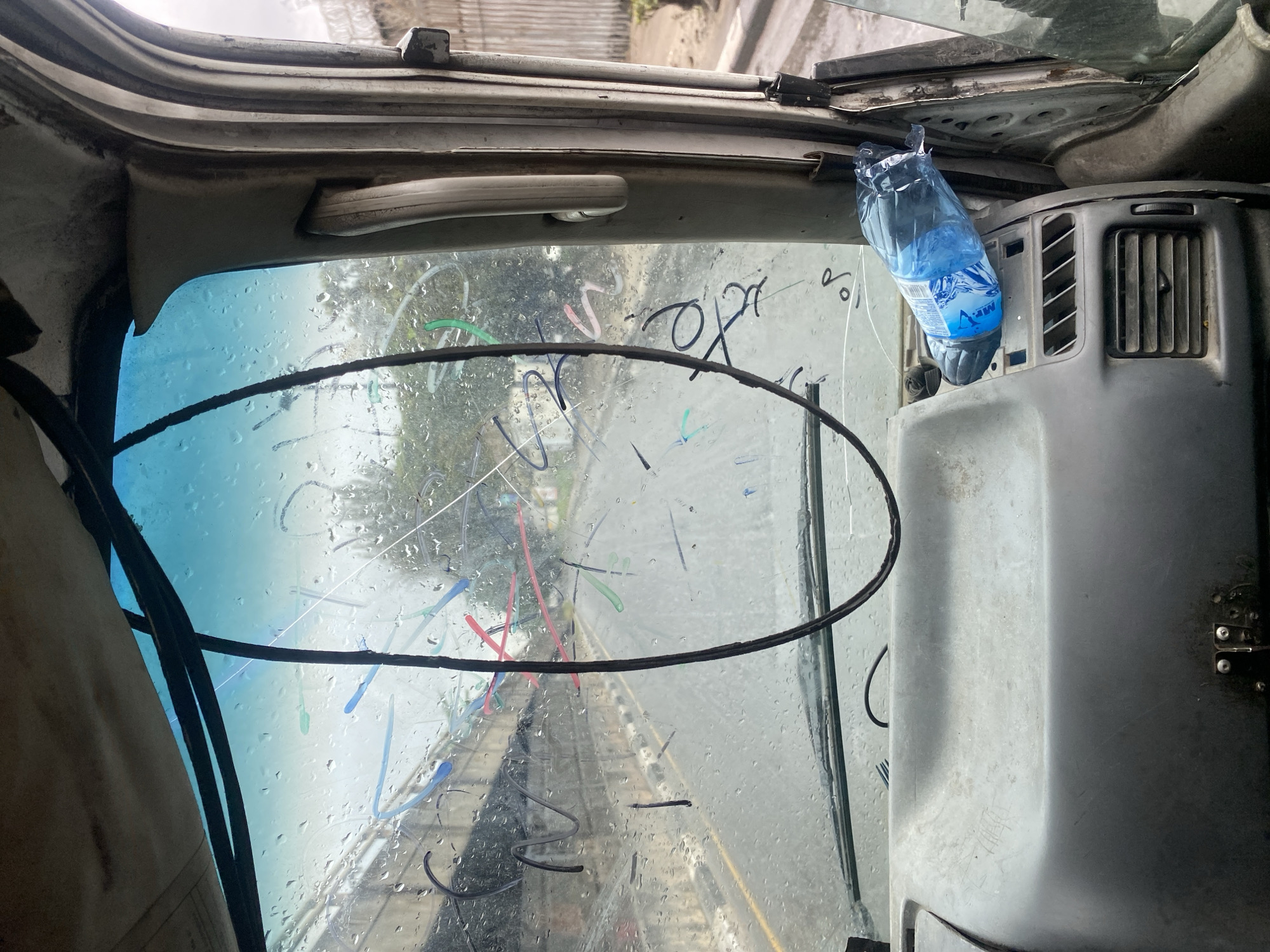 The image is taken from inside a vehicle, looking out through the windshield. The windshield is wet with raindrops and has various colorful scribbles on it, likely made with a marker. Outside, a road is visible, with a concrete structure on the left and trees and buildings in the distance. A plastic water bottle is placed on the dashboard to the right. The interior of the vehicle appears aged and somewhat worn. The setting is implied to be Victoria Island, Nigeria, suggesting an urban environment.