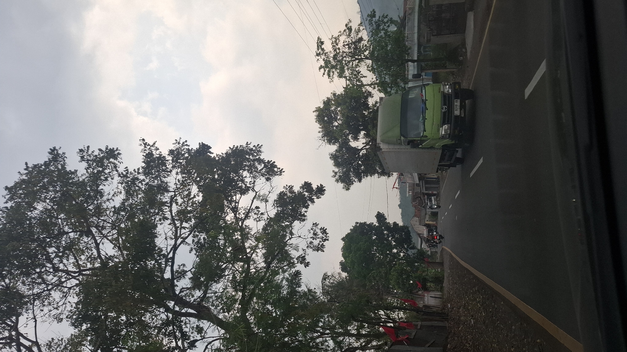 The image depicts a roadside scene in Susukan, Indonesia, likely captured from a vehicle. A green truck is the dominant subject, driving on a wet asphalt road. The truck, with a light-colored cargo box, has a license plate that reads "F 6098 HK". To the left of the road, there are trees and some buildings. Red flags are visible along the left side of the road, possibly indicating a celebration or event. Further down the road, other vehicles, including motorcycles, are present. The background shows more buildings and a hilly landscape under an overcast sky. The overall atmosphere suggests a slightly hazy, possibly early morning or late afternoon lighting.