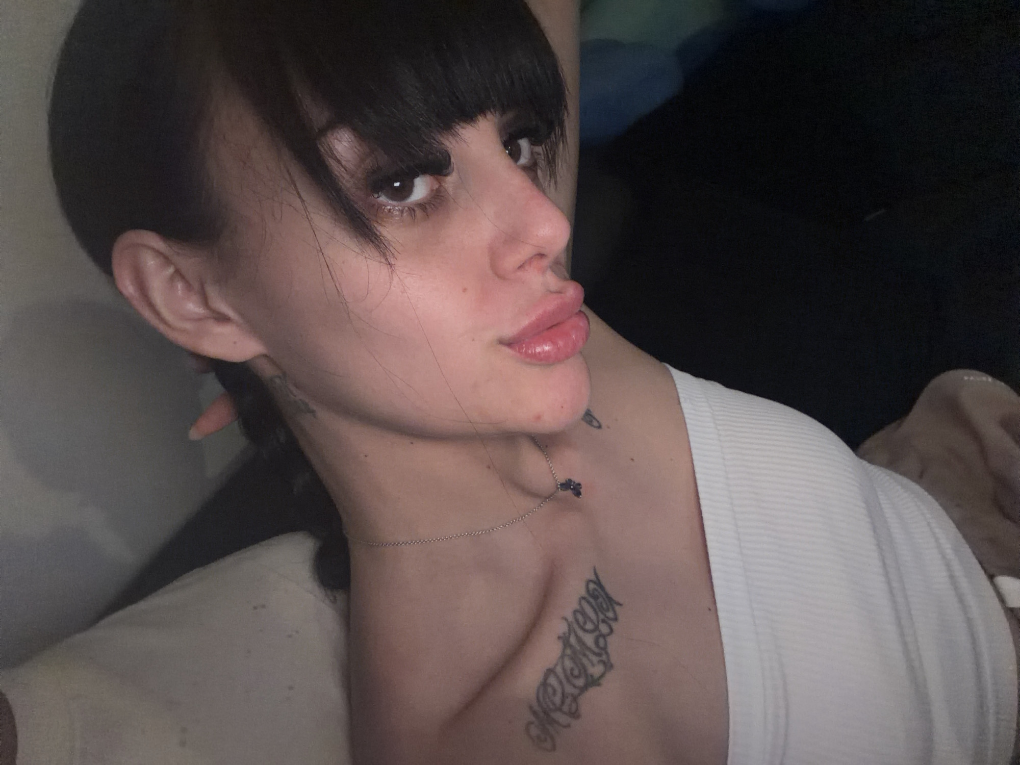 This is a close-up, indoor portrait of a young woman with dark hair and bangs. She is wearing a white ribbed tank top and a silver necklace with a small pendant. She has tattoos on her chest and neck. Her head is tilted to the side, and she is looking towards the camera with her lips slightly parted. The background is dark and out of focus, suggesting a dimly lit room. There is no visible information in the image that would indicate its location in Plovdiv, Bulgaria.