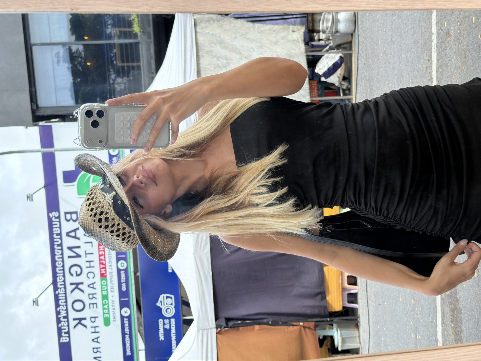 A woman wearing a black strapless dress and a straw cowboy hat takes a mirror selfie in front of a market in Choeng Thale, Thailand. She is holding a smartphone in her right hand. Behind her, market stalls are set up, with tents and various goods visible. A large billboard with Thai writing is also present in the background. The ground is paved, and there are some scattered leaves. The sky is overcast.