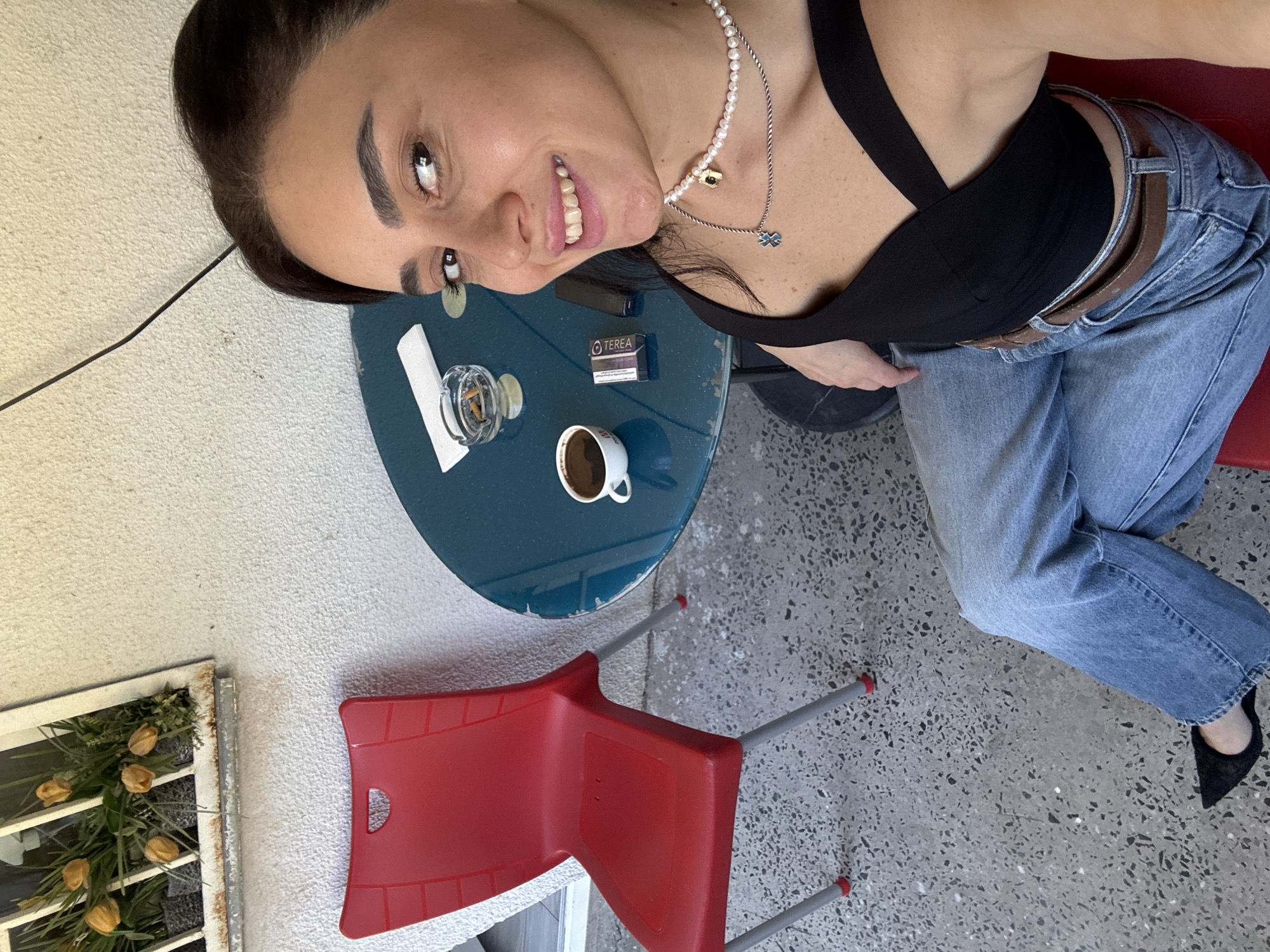 A woman is taking a selfie outdoors at a table with a cup of coffee, an ashtray with cigarettes, and a pack of Terea. She is wearing a black top, blue jeans, and black shoes. The table is blue and round, and a red chair is next to it. The ground is paved with grey concrete with black specks. In the background, there is a white wall and a window with yellow tulips. The scene is consistent with an outdoor cafe in Tbilisi, Georgia.