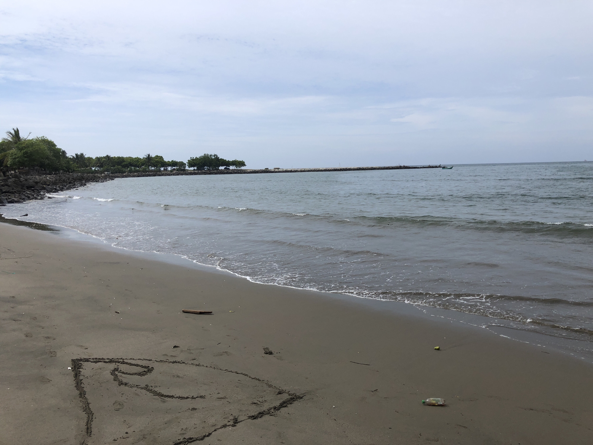 The image shows a wide, sandy beach with gentle waves lapping at the shore. The sand is a dark, wet color near the water and drier and lighter further up the beach. In the foreground, a heart shape has been drawn in the sand. A piece of driftwood lies on the sand to the left of the heart, and a plastic bottle is visible further to the right.
In the background, a line of rocks creates a breakwater, with trees and vegetation lining the coast behind it. The water extends to the horizon, meeting a cloudy sky. A small boat can be seen in the distance on the water, and a long, thin structure, possibly a pier or another breakwater, extends far out into the sea.
This scene is consistent with the appearance of the beaches in Lhokseumawe, Indonesia, a coastal city known for its proximity to the sea and its natural shorelines. Specifically, this could be a view of the Ujung Blang Beach or a similar coastal area within Lhokseumawe. There are no people visible in the image. The overall atmosphere is serene and natural, with a touch of human presence indicated by the drawing in the sand and the plastic bottle.