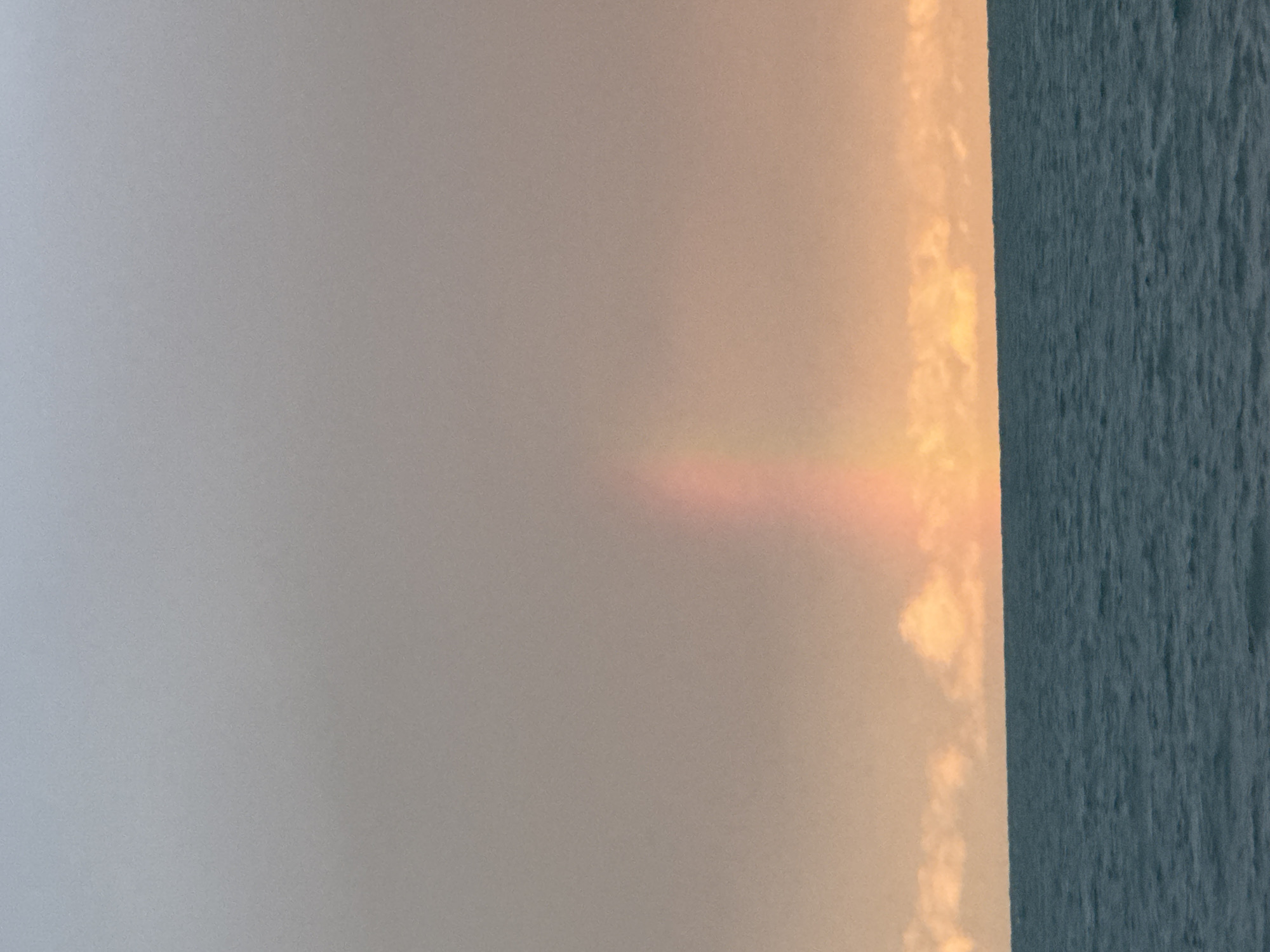 The image depicts a serene seascape with a faint rainbow arching across a hazy sky. The horizon line separates the deep blue, slightly rippled water from the sky, which transitions from a pale grey at the top to a warm, peachy hue near the water. A band of soft, illuminated clouds stretches across the sky just above the water's edge. In the center of the frame, a subtle, vertical arc of pastel colors suggests the presence of a rainbow, though it is not fully formed or intensely vibrant. The overall impression is one of tranquility and natural beauty. The location is described as Ақши, Kazakhstan, which is a town situated on the banks of the Kapchagay Reservoir, a large artificial lake in the Almaty Region of southeastern Kazakhstan.