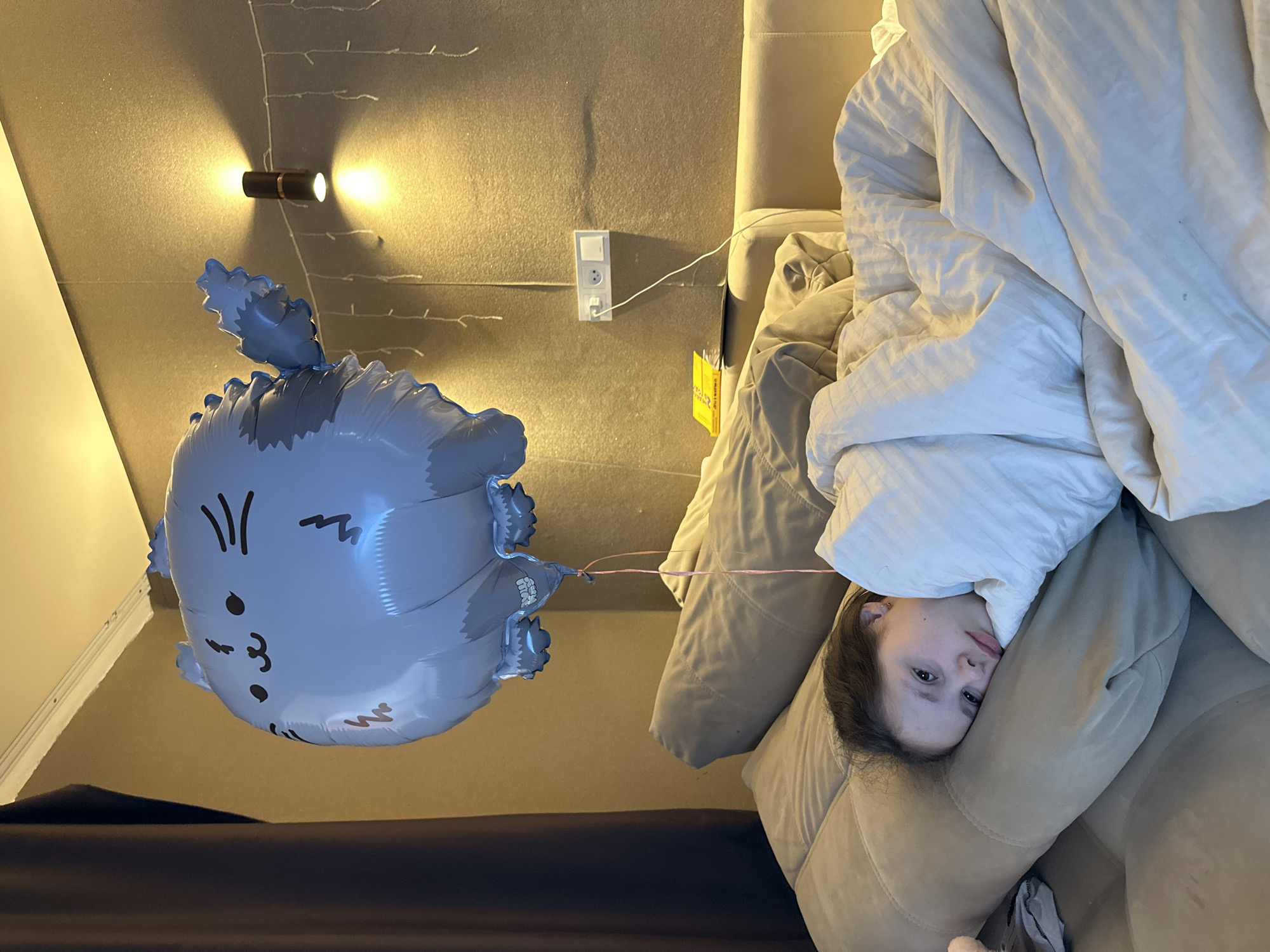 A young person is lying in bed with a gray cat-shaped balloon nearby. The room has a wall light and an electrical outlet. The location is Unknown, Unknown.