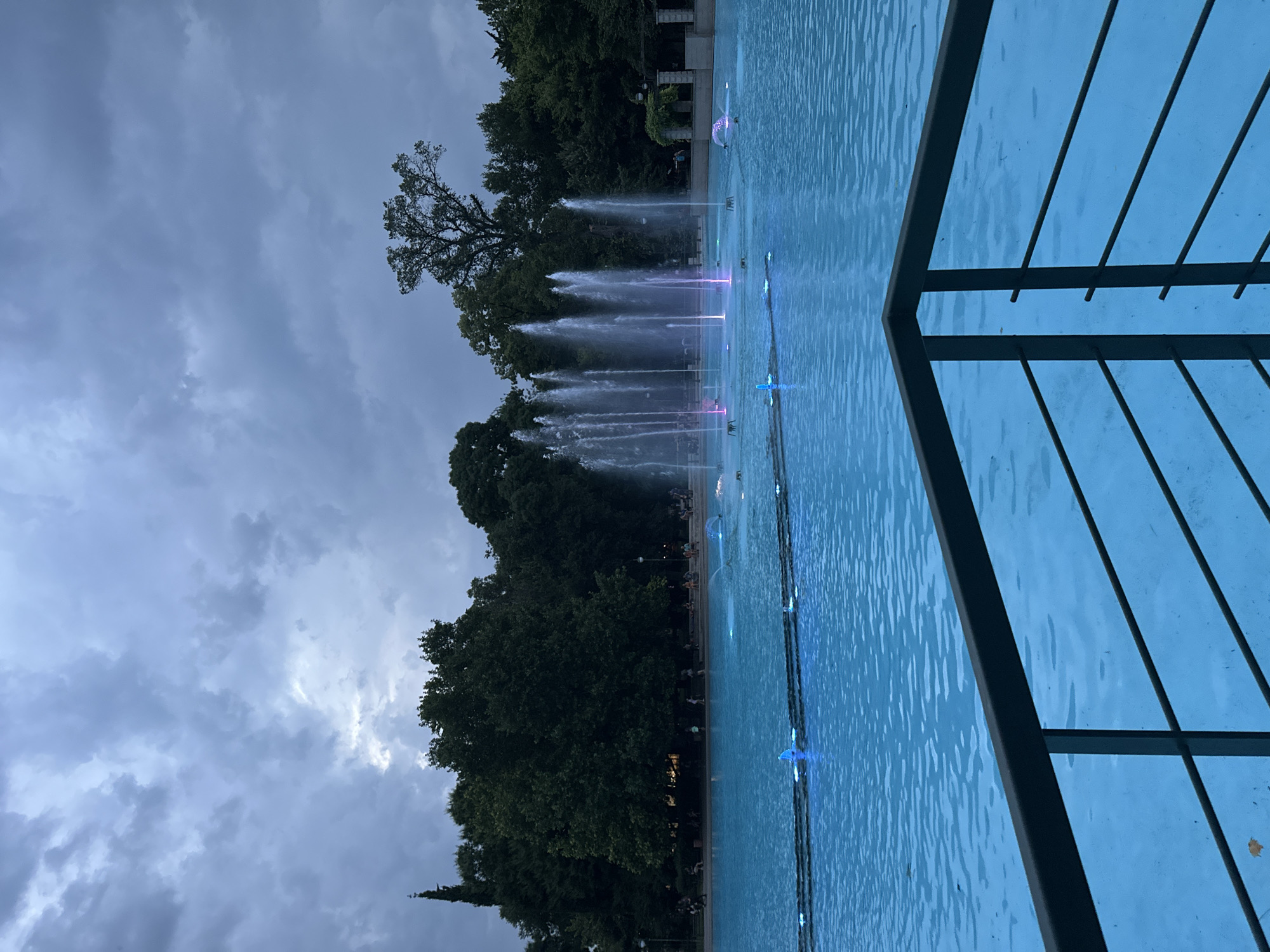 The image shows a large, brightly lit blue fountain in a park setting. The fountain has multiple water jets, some of which are illuminated with pink and purple lights. The surrounding area is filled with lush green trees, and a cloudy sky looms overhead. In the foreground, a dark metal railing divides the viewer from the fountain. A few people can be seen in the background, walking and sitting near the fountain. This scene depicts the Singing Fountains of Plovdiv, Bulgaria, which are located in the Tsentralen Park.
