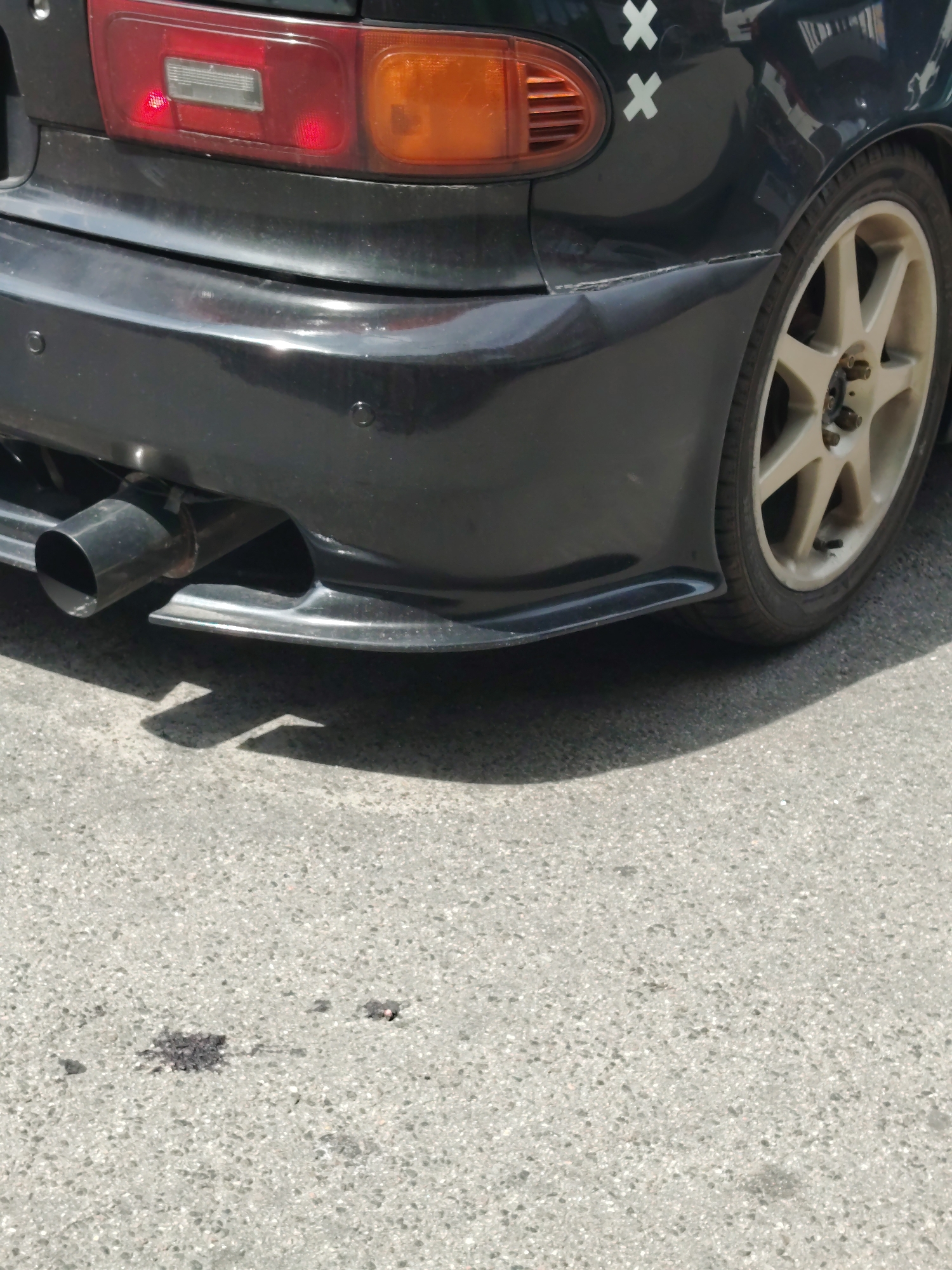 A black car is parked on asphalt. The car's rear bumper is visible, along with a portion of the taillight and a wheel. The car has two white 'X' decals on its fender. There are dark stains on the asphalt in front of the car, possibly oil or another fluid. The environment appears to be an outdoor urban setting.