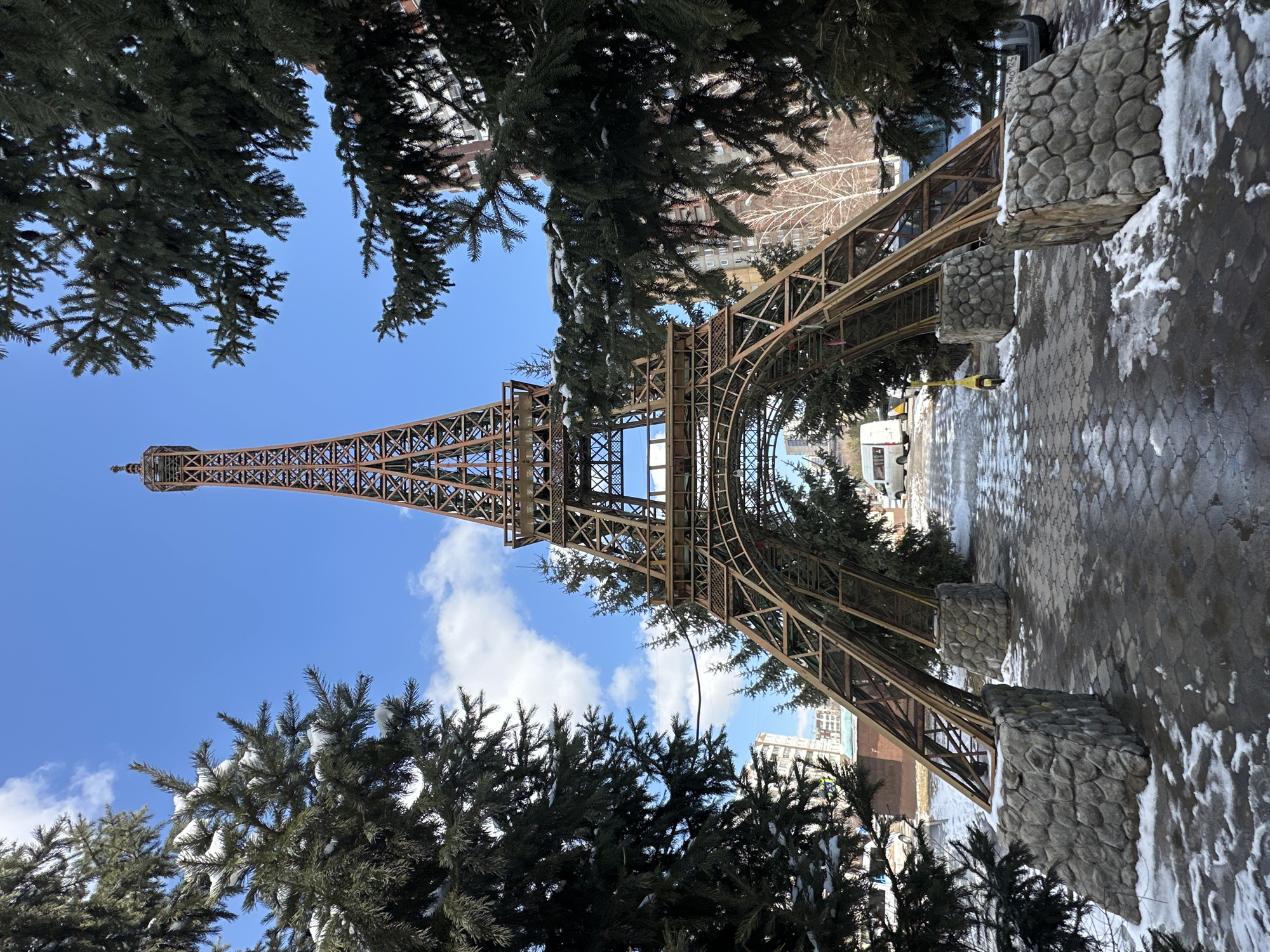 A low-angle shot depicts an Eiffel Tower replica in Krasnoyarsk, Russia. The replica is framed by evergreen trees with patches of snow on their branches. The sky is blue with scattered clouds. The base of the Eiffel Tower replica is surrounded by pavement speckled with snow. The tower is made of brown metal. Buildings can be seen in the background.