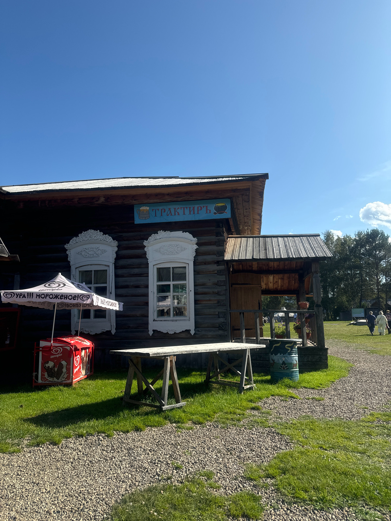A rustic wooden building, identified by a sign as "ТРАКТИРЪ" (Trakir), stands under a clear blue sky in the Museum of Wooden Architecture in Taltsy, Russia. The building has two windows with decorative white carved surrounds. To the left of the building is a white umbrella advertising "УРА!!! МОРОЖЕНОЕ!" (Hooray!!! Ice cream!) over a red ice cream freezer. A wooden table with X-shaped legs is set up in front of the building, casting a shadow on the green grass. In the background, several people are walking on a gravel path, and trees form a dense line at the edge of the property. The overall scene depicts an outdoor setting, likely a historical or cultural park, with elements suggesting a place of business or refreshment.