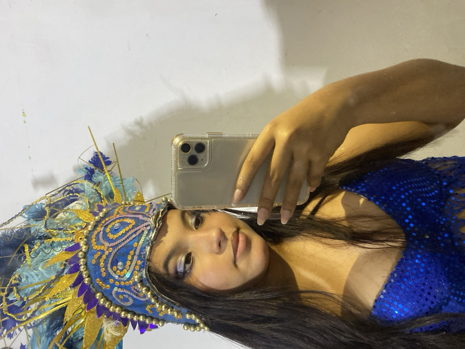 A person poses for a selfie, holding a phone up to their face. They are wearing a large, ornate blue and gold feathered headdress. Their outfit appears to be a matching blue sequined garment. The background is a plain white wall. The photograph is rotated, with the person's head tilted to the side.
