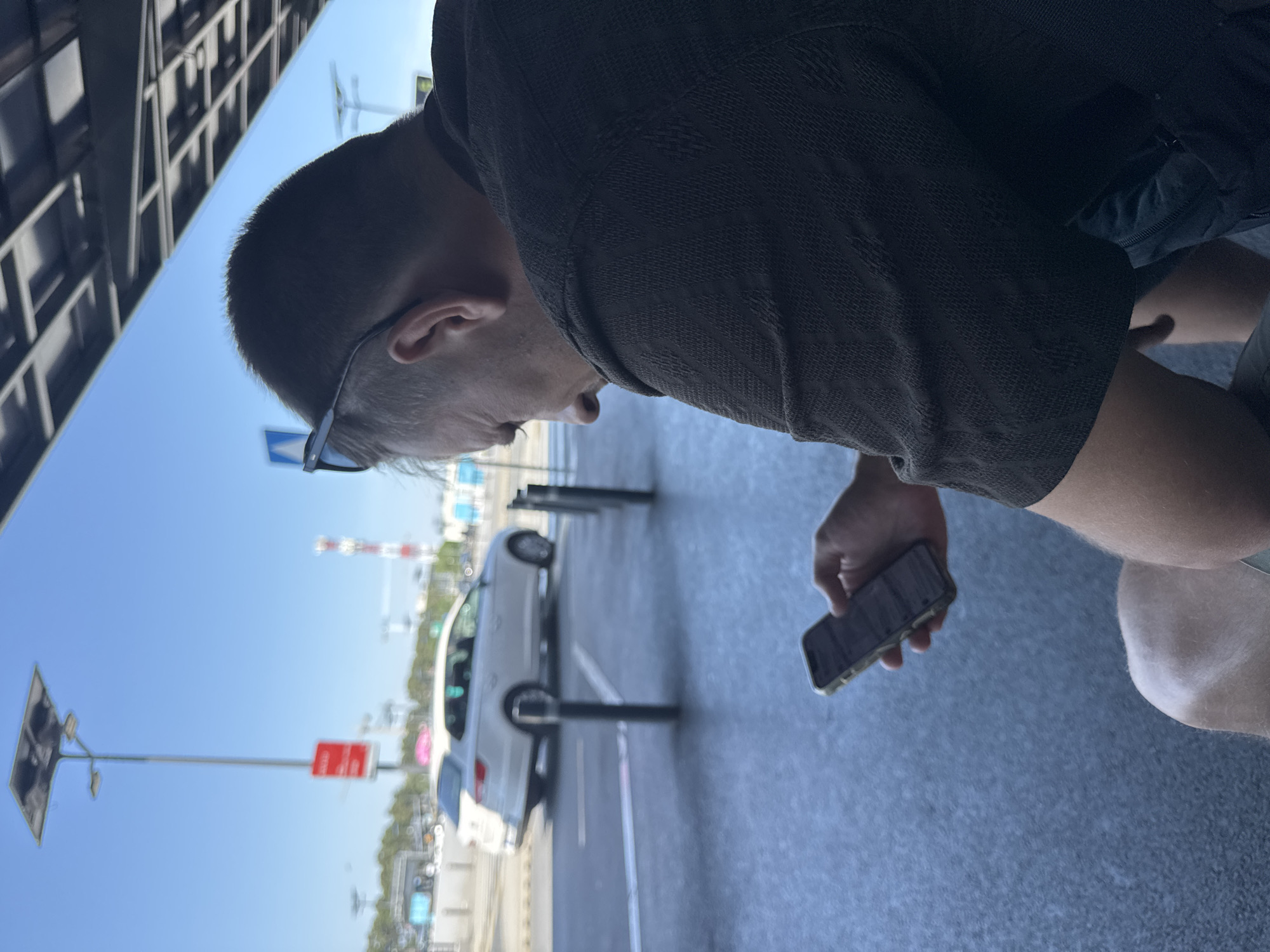 A man is sitting outdoors, holding a smartphone in his right hand and looking at it. He is wearing sunglasses pushed up on his head and a dark patterned shirt. The background shows a bright blue sky, a white station wagon driving on a wet asphalt road, and a tall street light with solar panels on top. The location appears to be an outdoor public space, possibly an airport or transportation hub, given the signage and infrastructure visible in the background. The overall lighting suggests a sunny day.