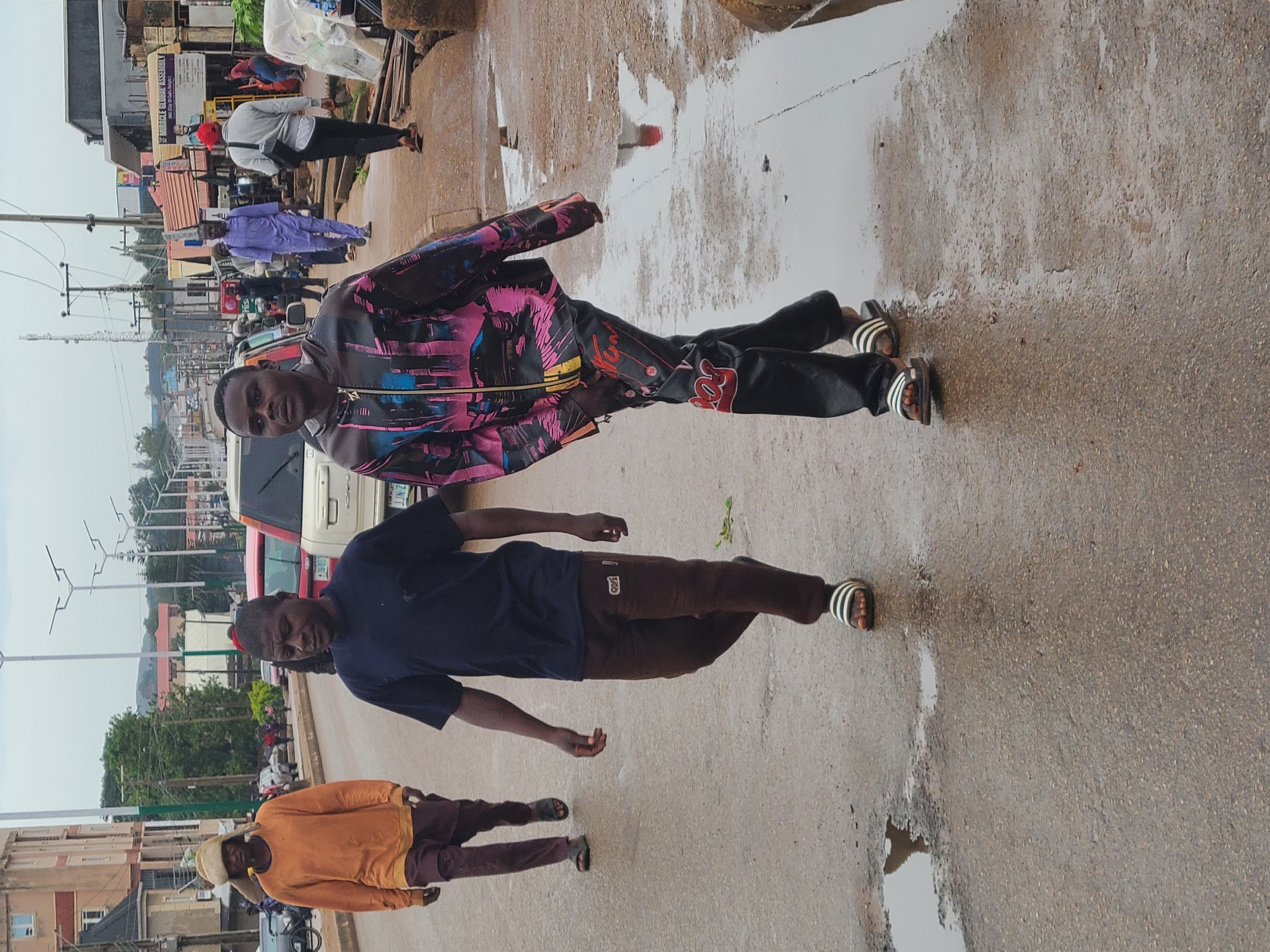 The image shows a street scene in Unknown, Unknown. Three men are visible, one wearing a black shirt and brown pants, another in a colorful jacket and black pants, and a third in an orange shirt and brown pants. The ground appears wet with puddles. In the background, there are buildings, cars, and streetlights.