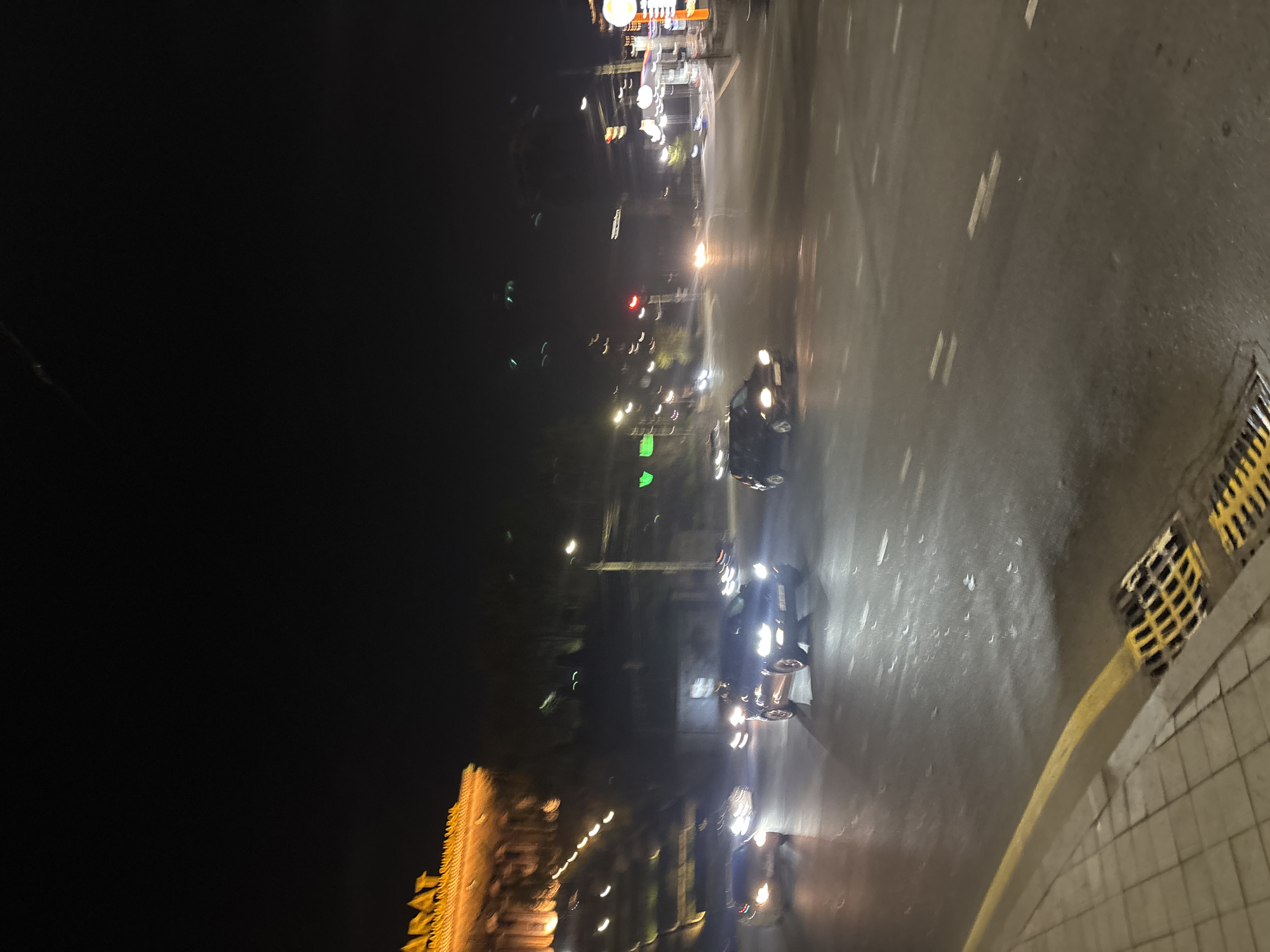 The image depicts a street scene in Yerevan, Armenia at night. The street is wet, possibly from rain. Cars are driving on the road, their headlights illuminating the surface. Streetlights and building lights provide additional illumination. On the right side of the image, there is a sidewalk with a yellow curb and a drainage grate. In the distance, illuminated buildings can be seen. The image is somewhat blurry, suggesting movement during the photo capture.