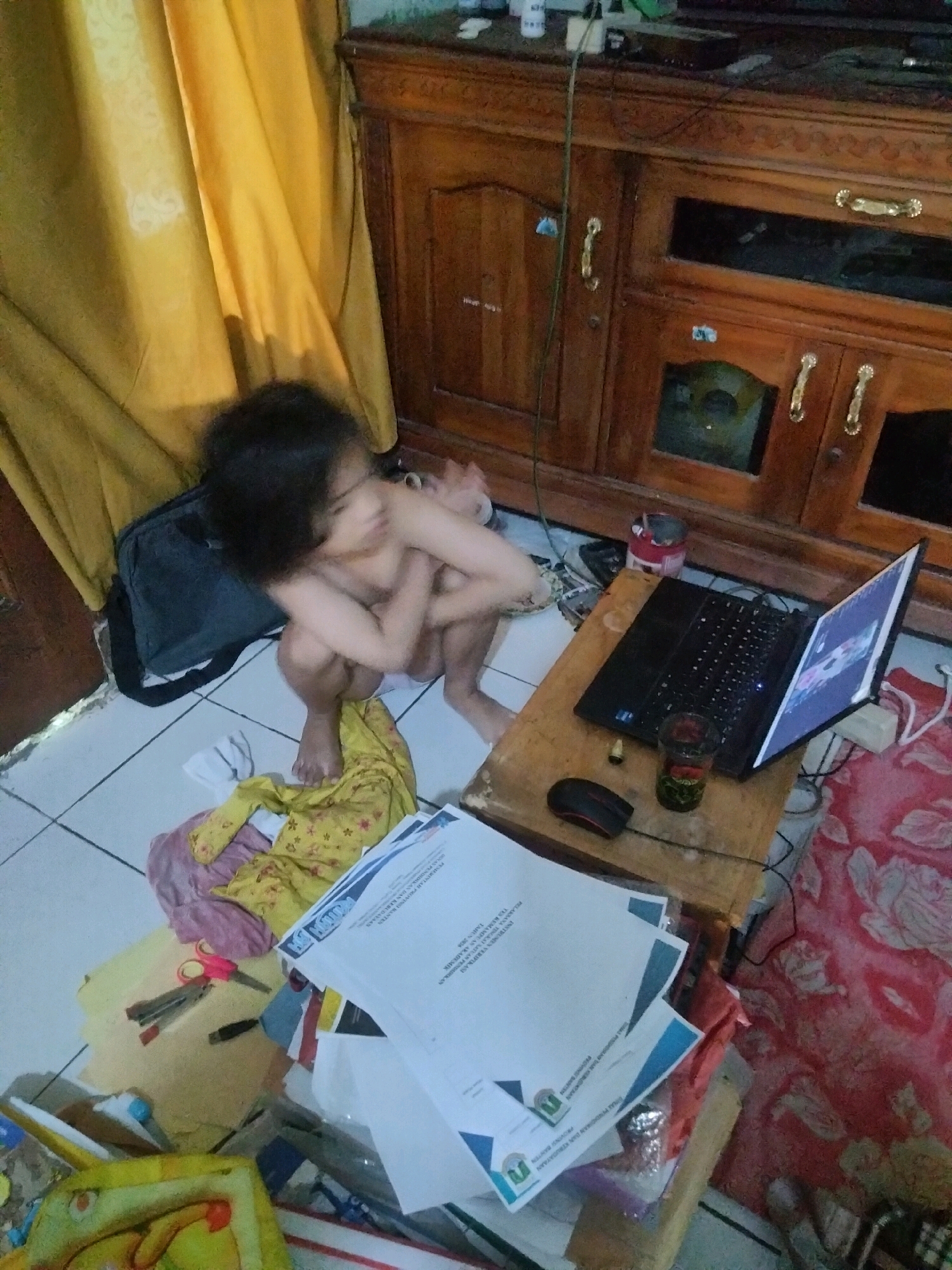 This is a medium shot of a young child in a squatting position in what appears to be a bedroom or living area. The child is unclothed and looking towards the right of the frame. The child's dark hair is tousled. In the background, a large wooden cabinet with glass-fronted doors is visible. To the left, a yellow curtain is drawn.
In the foreground, a small wooden table holds a laptop, a computer mouse, and a glass with liquid. Various papers are stacked on a lower surface, along with a pair of scissors and a stapler. A patterned cloth and a black bag are also on the floor. The floor is tiled. The scene suggests a home environment, likely in the daytime given the lighting from the yellow curtain. There is some visible text on the papers, including "DINAS PENDIDIKAN DAN KEBUDAYAAN PROVINSI BANTEN" and other Indonesian phrases related to academics and verification. The overall impression is one of a somewhat cluttered but lived-in space.