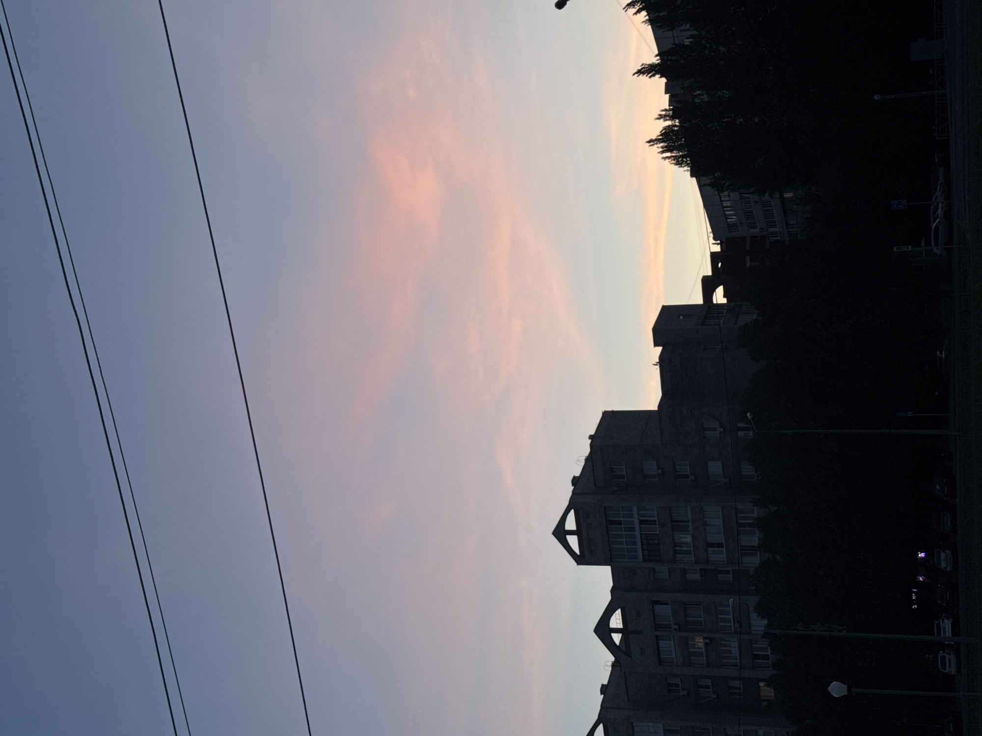 The image shows a view of an urban landscape at dusk or dawn. The sky is a mix of pale blue and pink hues, suggesting either a sunset or sunrise. In the foreground, there are two power lines running diagonally across the frame. Buildings, mostly residential apartments, are visible in the middle ground. The buildings are dark and gray, obscuring architectural details. Dark silhouettes of trees line the street, adding to the overall contrast. The image is taken in the city of Unknown, Unknown.