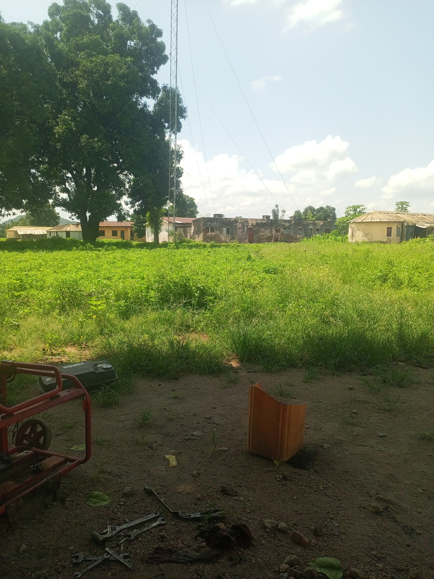 In Serti, Nigeria, an outdoor scene unfolds under a partly cloudy sky. In the foreground, a red metal generator with a black fuel tank sits on a dirt ground, accompanied by a collection of wrenches. To the right of the generator, an orange plastic container lies on its side. Behind this, a vast field of lush green vegetation stretches into the midground. Several buildings are visible in the background, including a long, dilapidated structure with visible windows and a single-story building with a light-colored roof. A tall metal tower rises behind the buildings, with thin wires extending from it. Large trees with dense green foliage are scattered throughout the scene, with the largest tree dominating the left side of the image. The ground is a mix of dirt, small stones, and patches of grass.